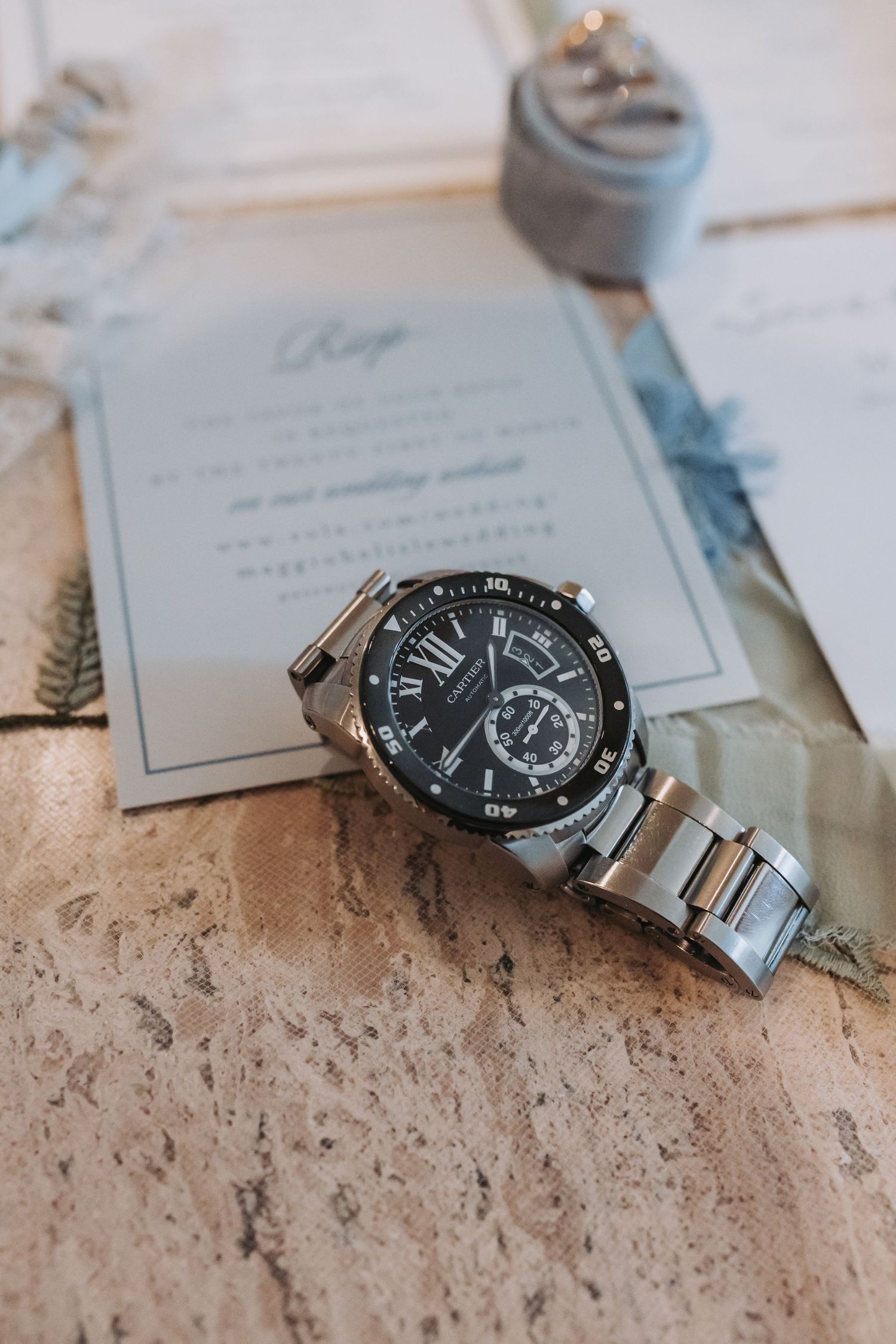Silver Cartier Calibre watch resting on wedding invitation stationery with blue ribbon and ring box on wooden surface.
