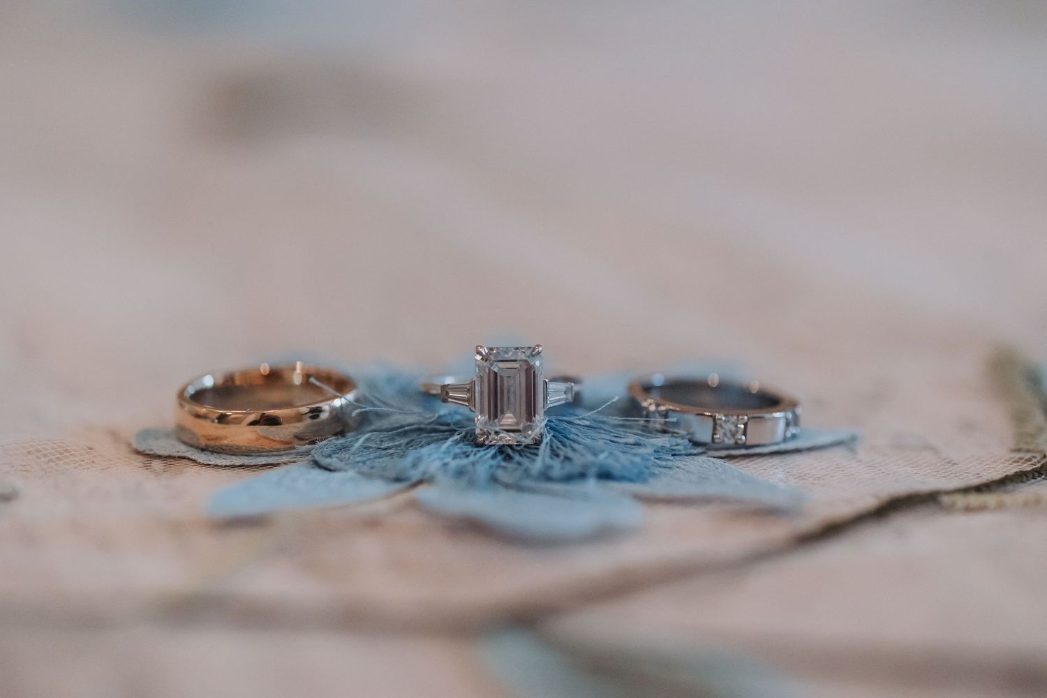 Three wedding rings displayed on blue fabric, featuring a rose gold band, emerald cut diamond, and diamond eternity band.