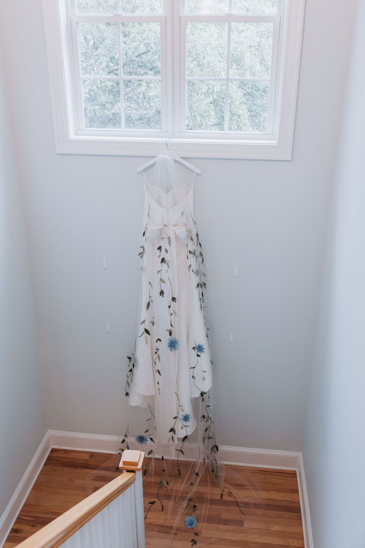White floral embroidered wedding dress hanging in a stairwell window with blue flower details.