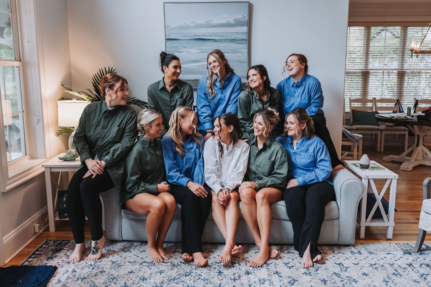 Bride poses with bridesmaids in matching green and blue jackets on a white couch before the wedding.