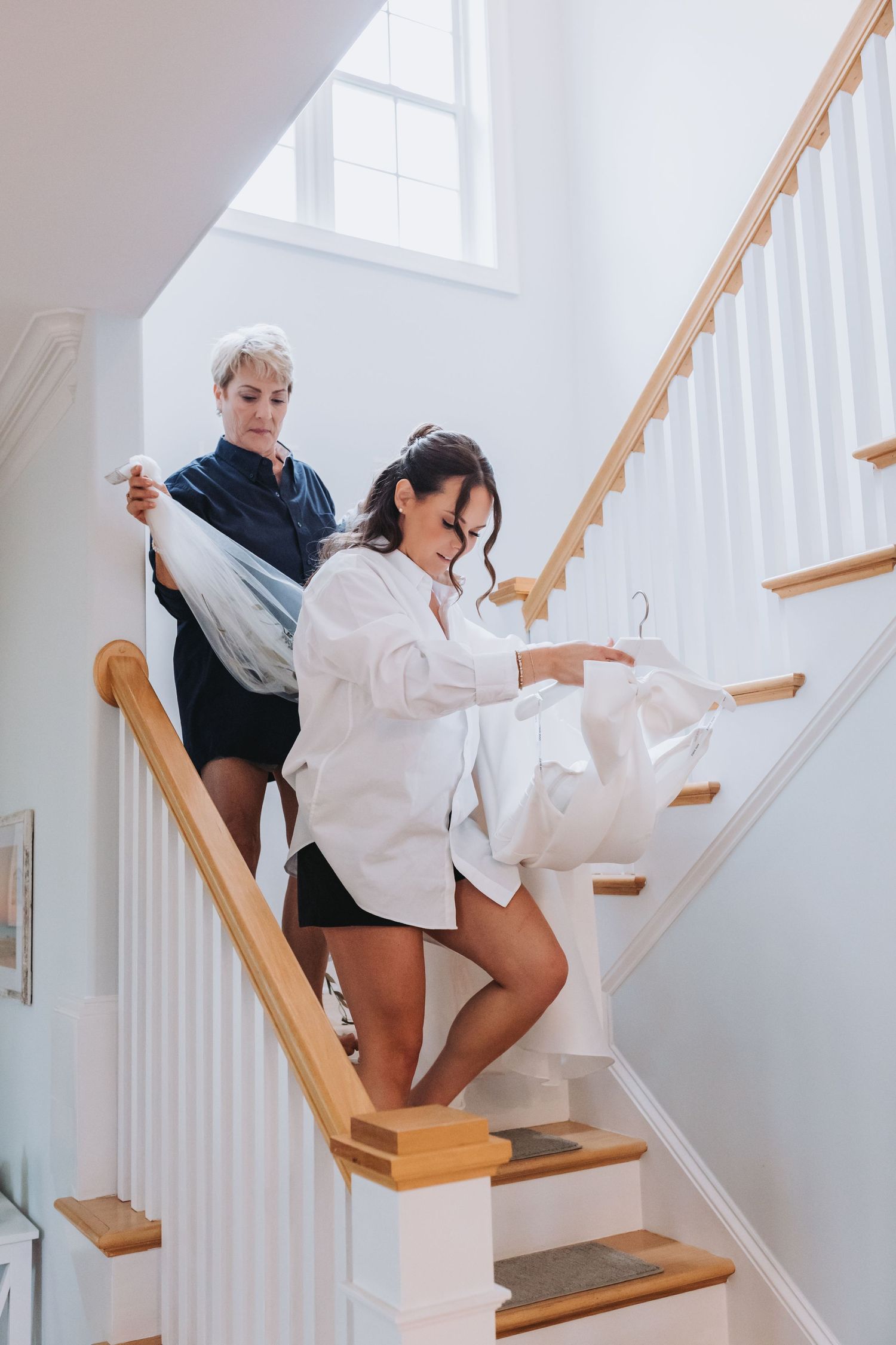 Bride in white button-down shirt carrying wedding gown on hanger down wooden staircase with assistant.