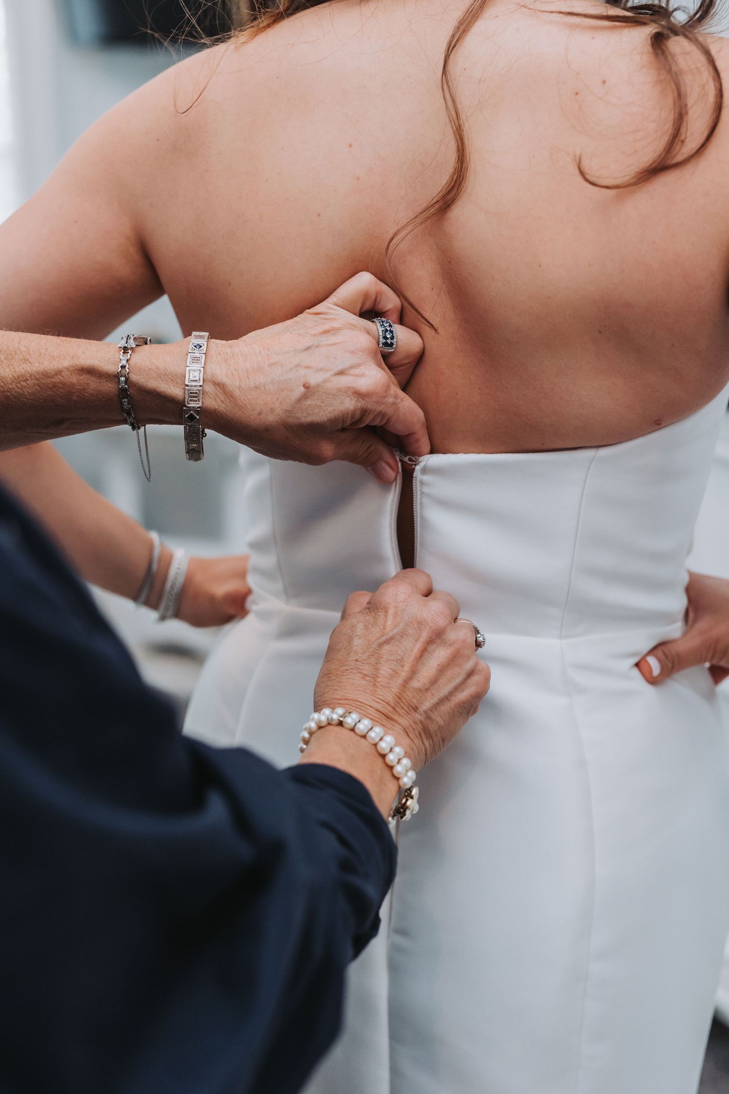 Two women with pearl and silver bracelets fasten buttons on back of brides white strapless wedding gown.