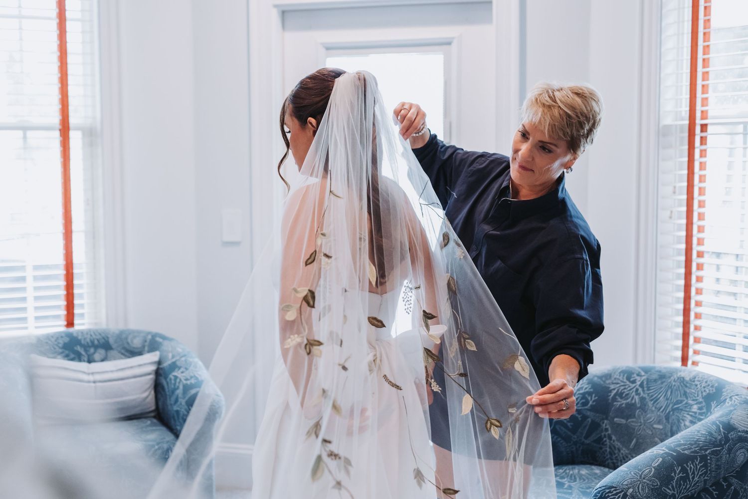 Wedding assistant carefully arranges bride's embroidered veil in bright dressing room before ceremony.