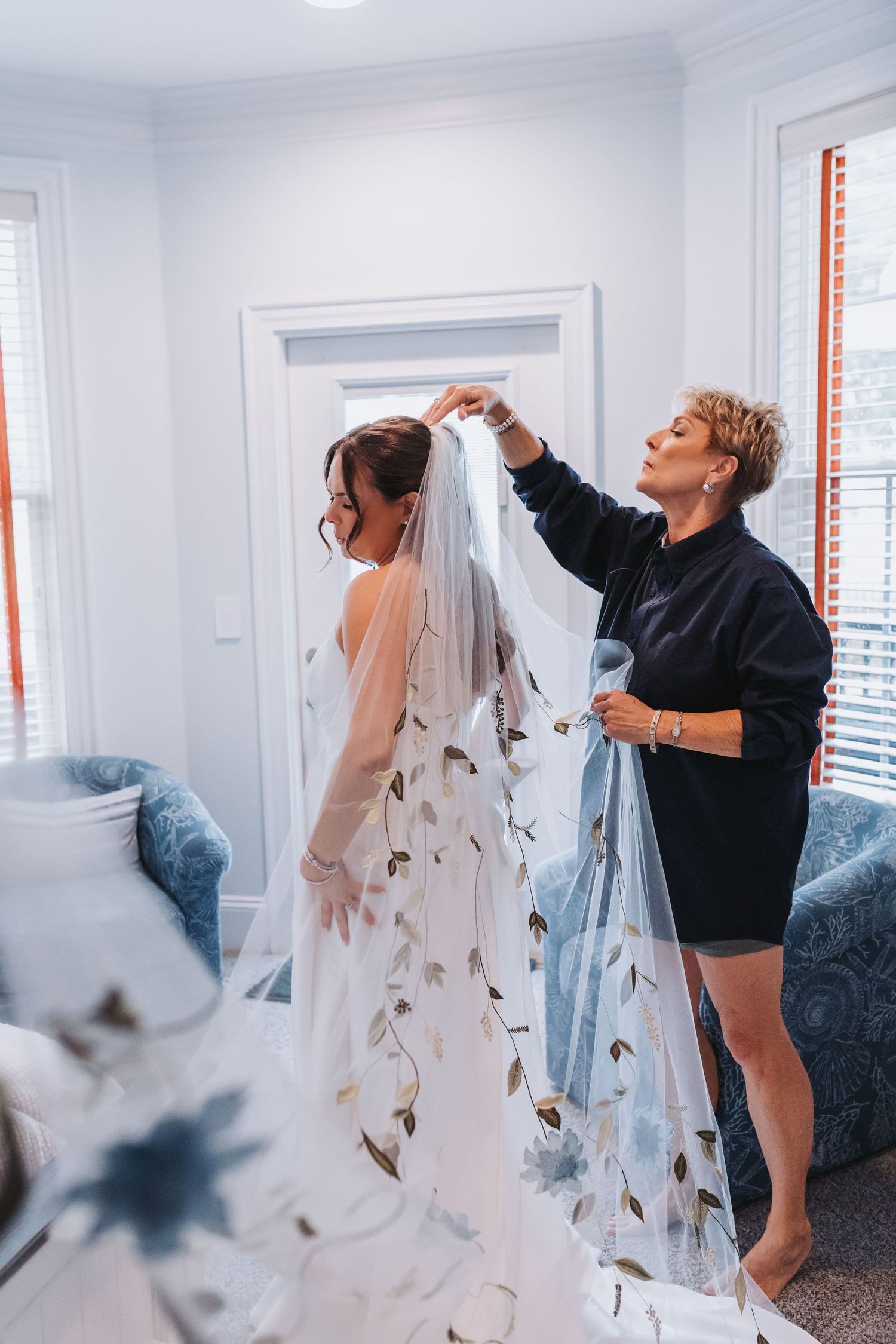 Woman helping bride attach embroidered floral veil to wedding dress in a bright, blue-accented dressing room.