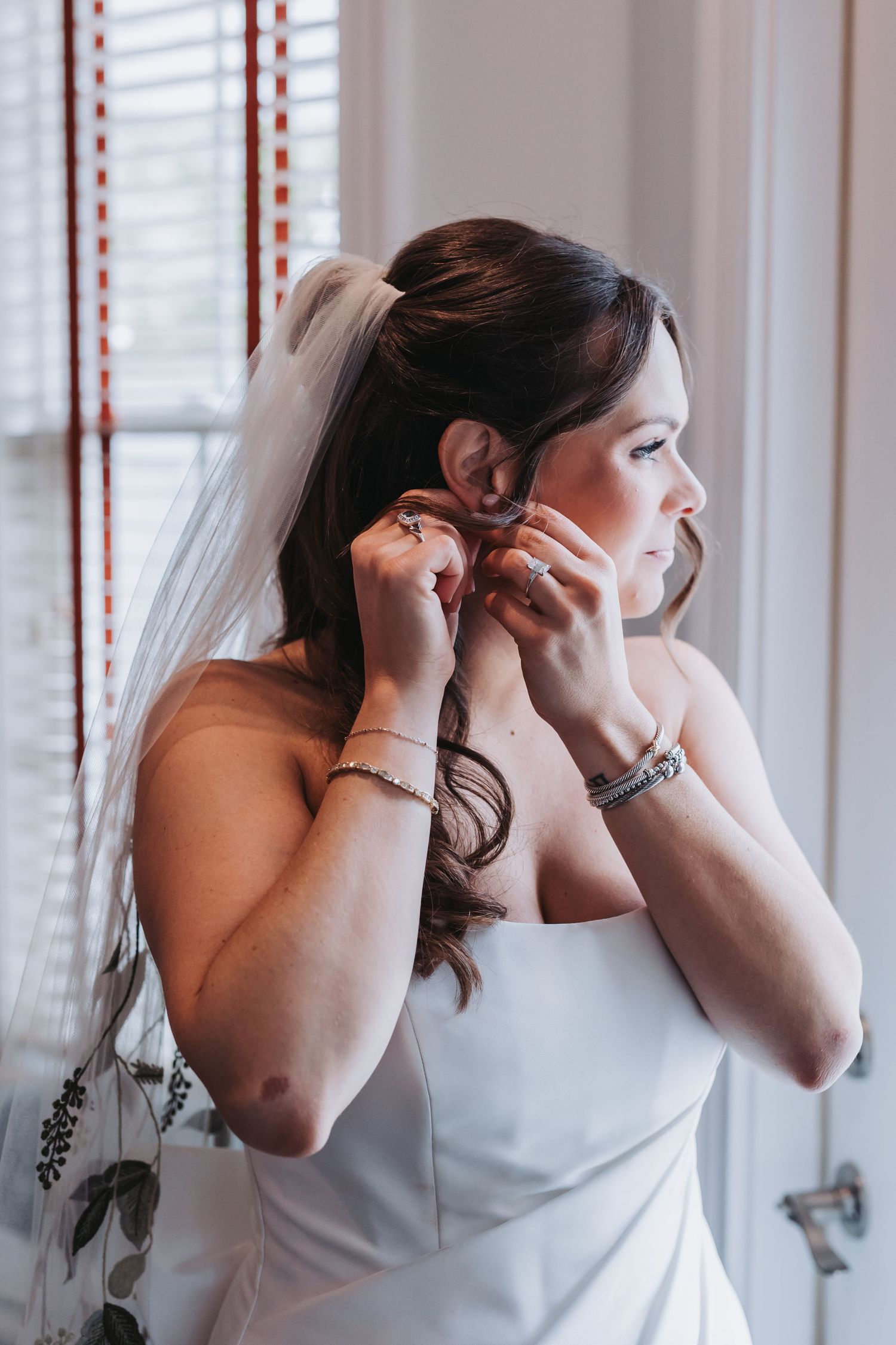 Bride in strapless white gown putting on earrings by window while wearing veil and diamond bracelet.