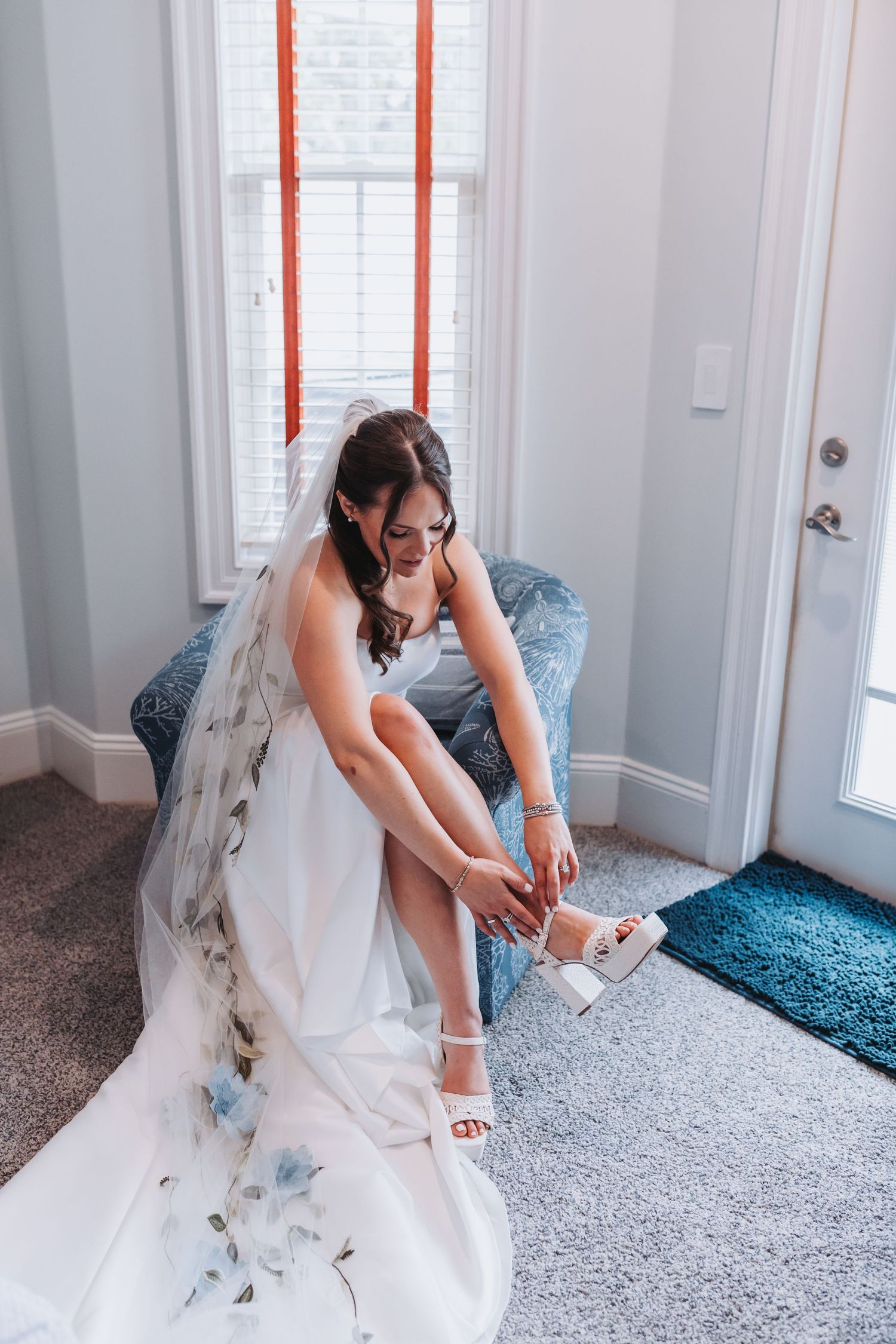Bride in floral wedding gown putting on white heeled sandals while seated in a blue chair.