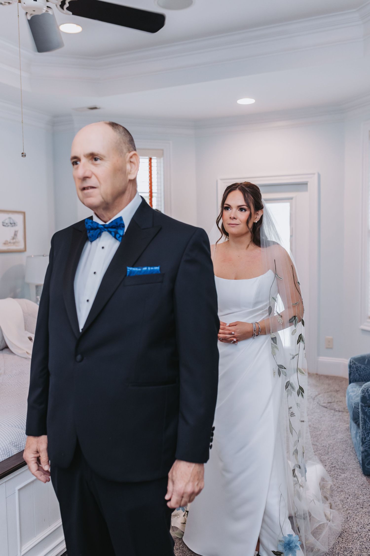 Bride in white strapless gown approaches father in black tuxedo with blue bow tie for emotional first look moment.