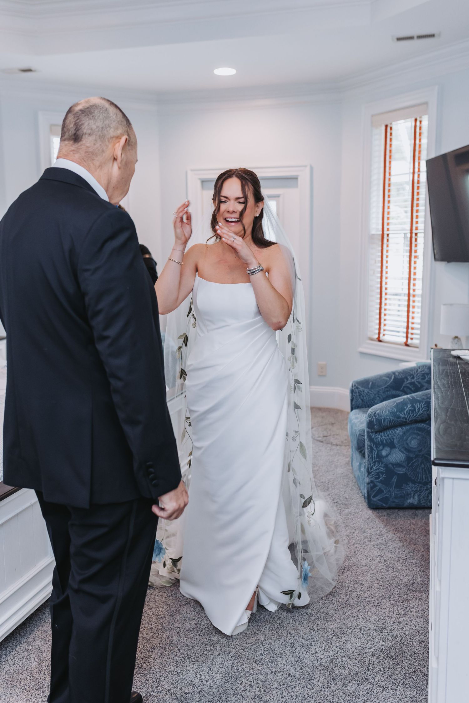 Bride in white strapless gown laughing with suited man during emotional first look moment indoors.