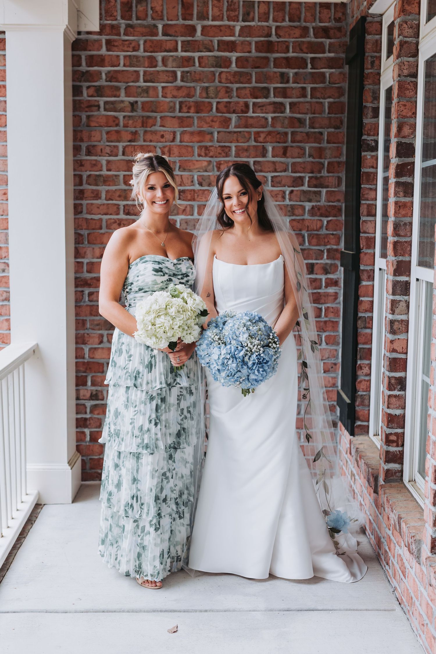 Bride in white gown and bridesmaid in floral green dress holding bouquets against brick wall backdrop.