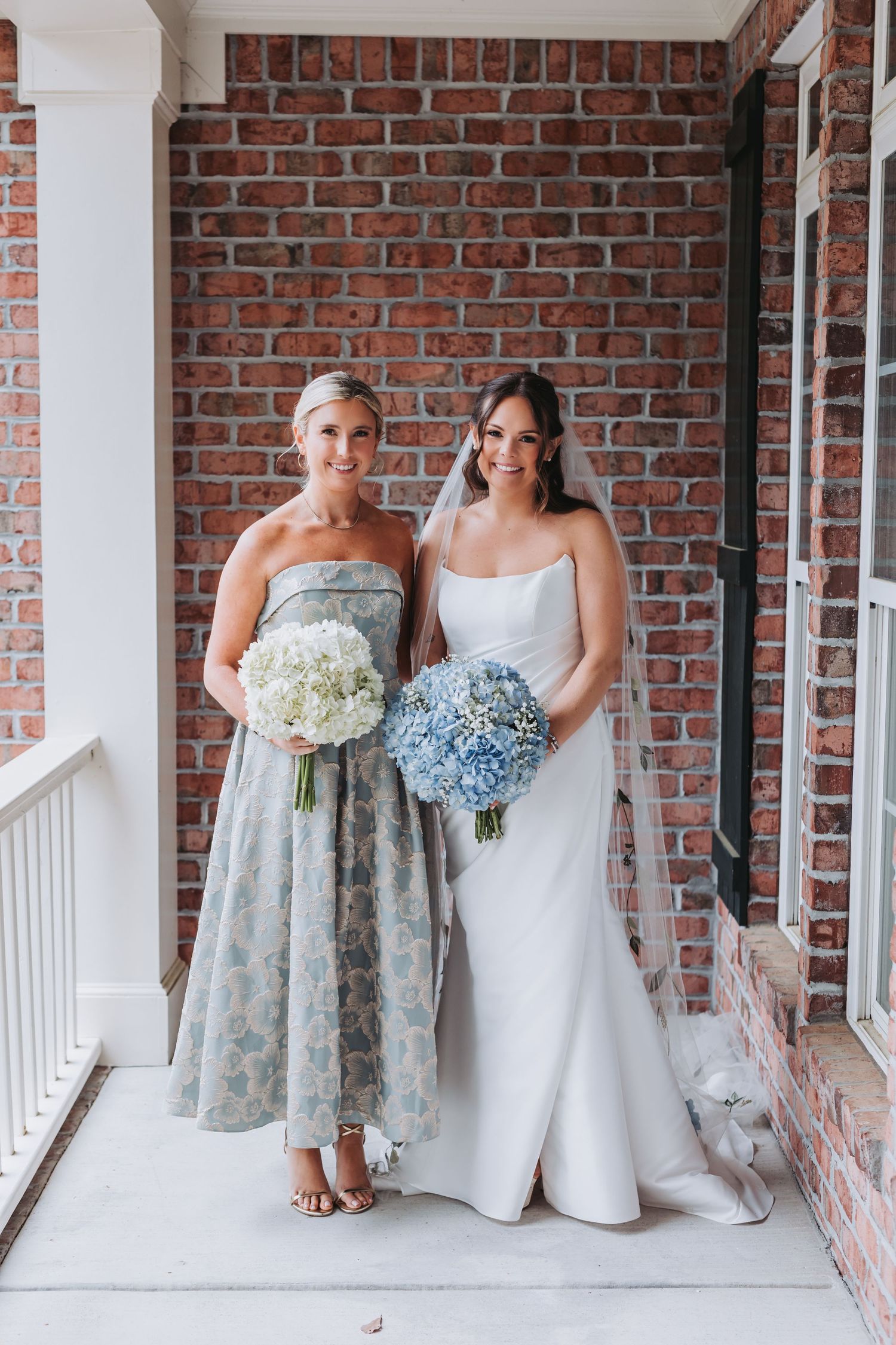 Bride in white strapless gown and bridesmaid in blue floral dress hold hydrangea bouquets against brick wall.