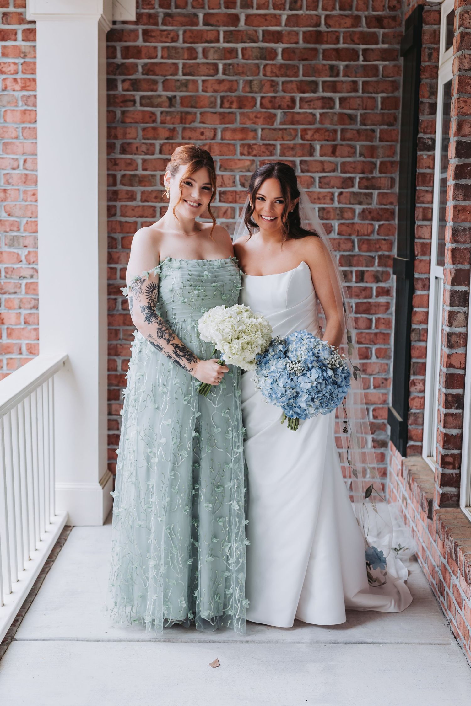 Bride in white dress and bridesmaid in sage green gown holding bouquets against brick wall.