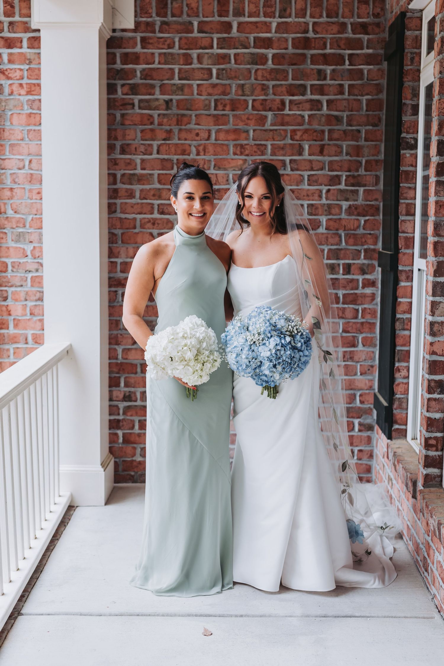 Bride and bridesmaid in sage halter dress posing with white and blue hydrangea bouquets on porch.