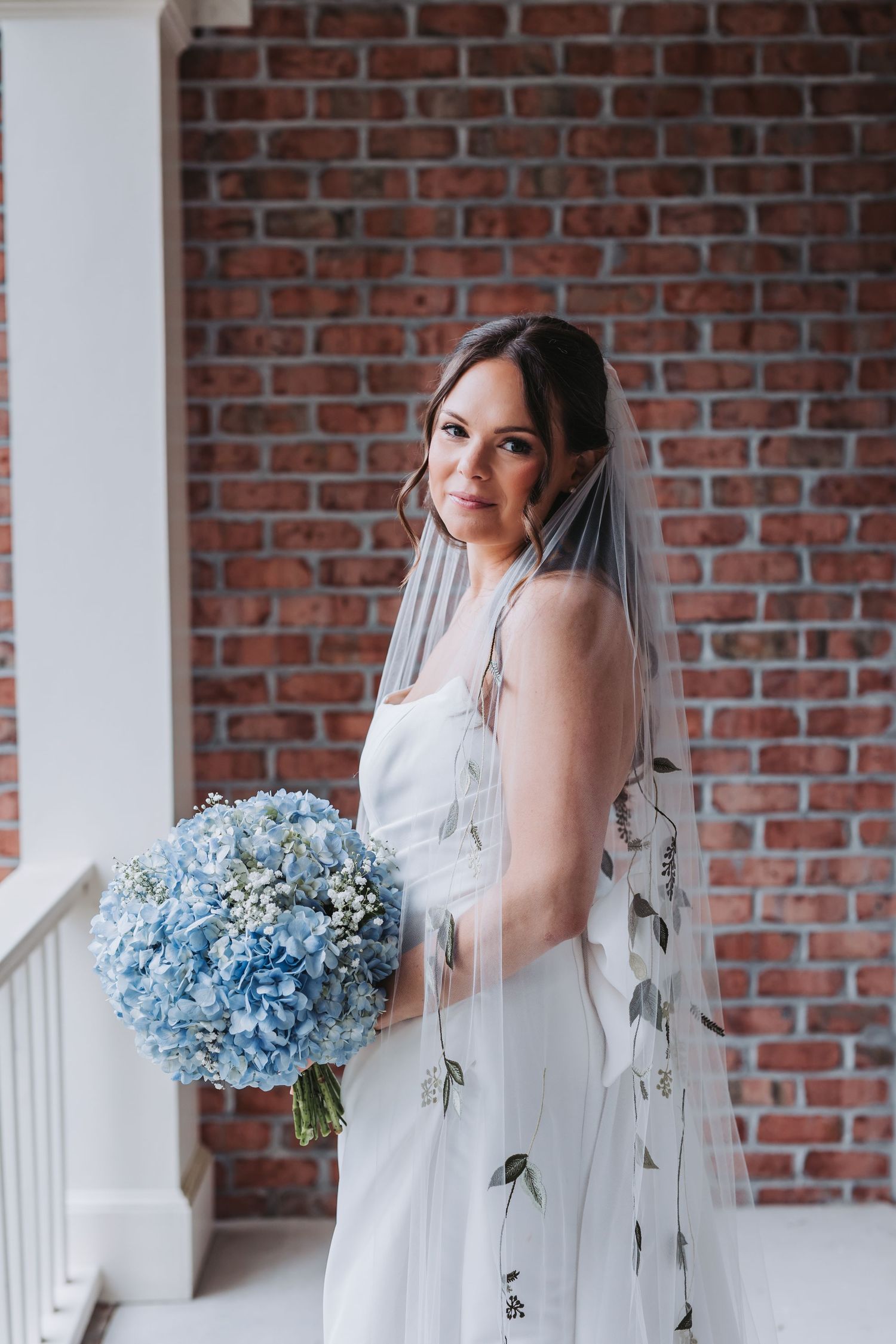 Elegant bride in white dress holding blue hydrangea bouquet wearing floral embroidered veil.
