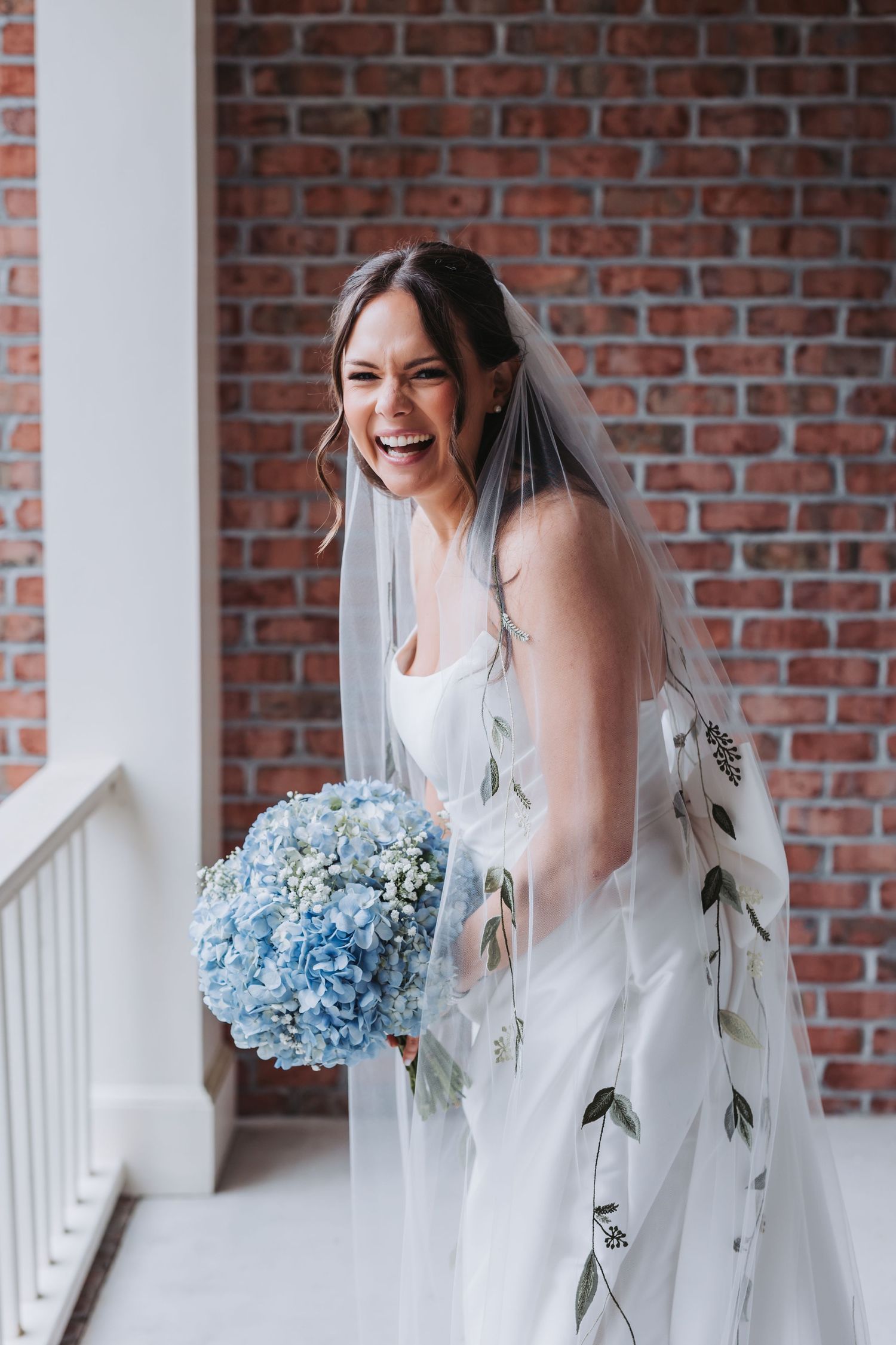 Joyful bride laughing in white gown and floral veil holding a blue hydrangea bouquet against a brick wall.