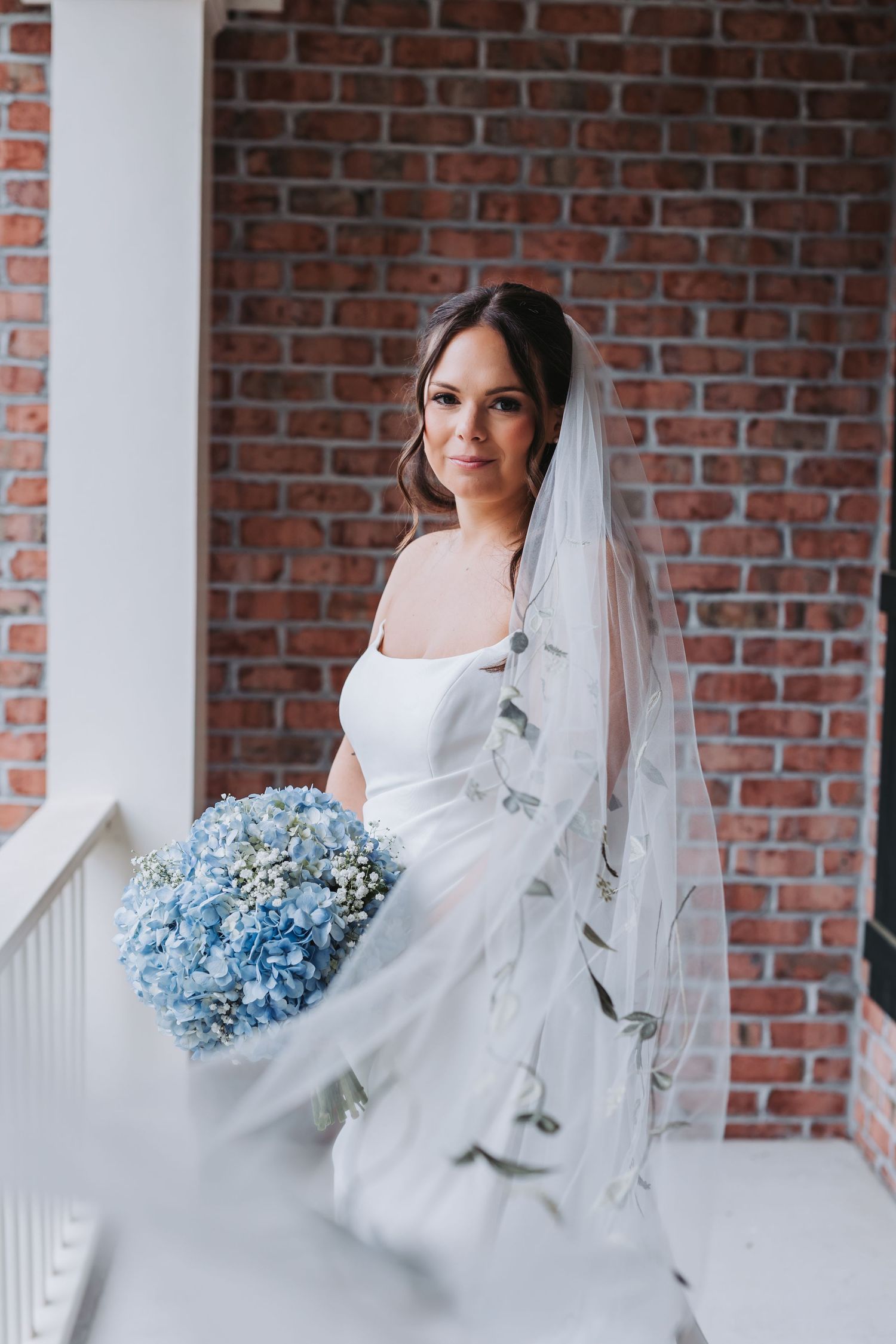 Bride in strapless white gown with embroidered veil holding blue hydrangea bouquet on porch.