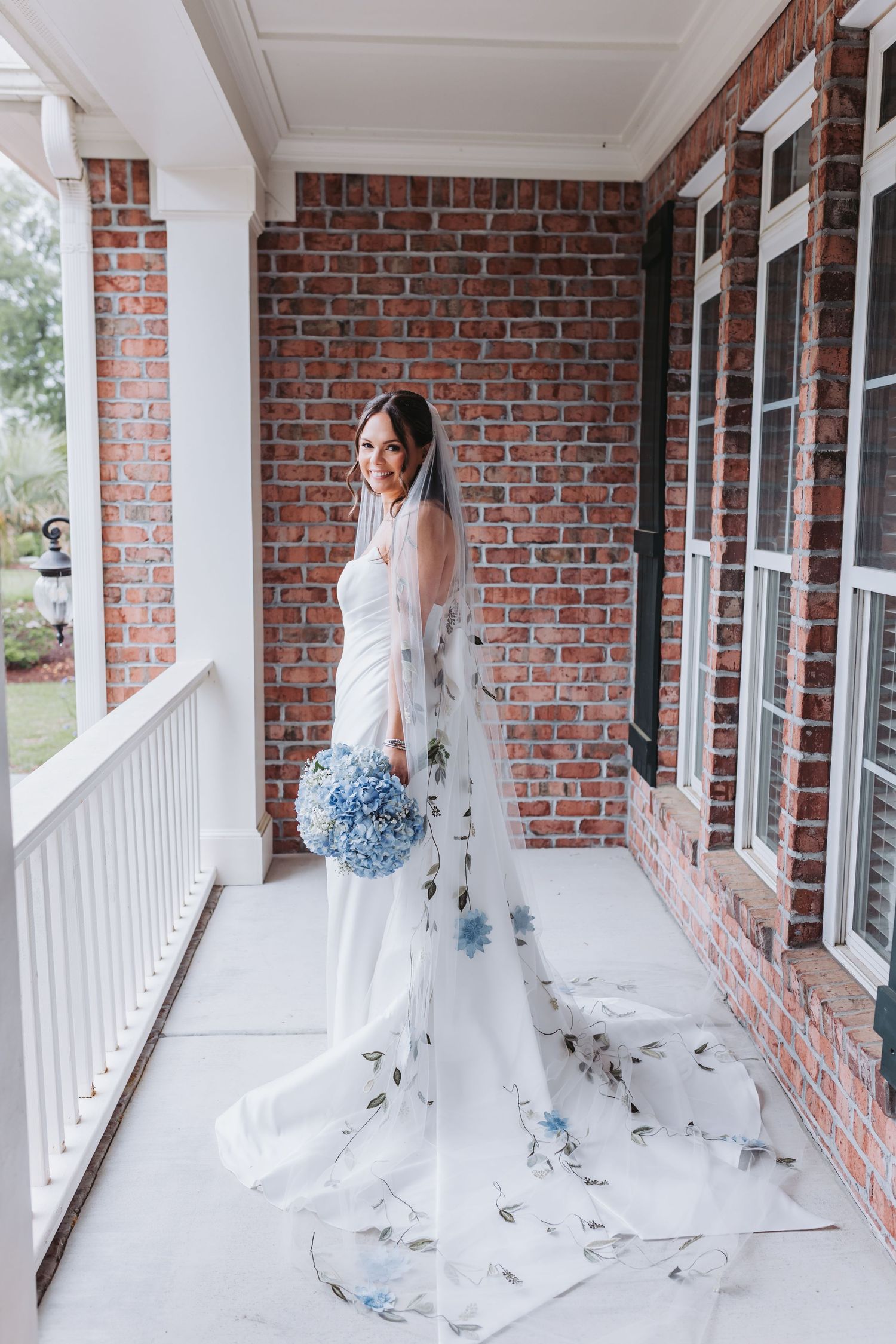 Elegant bride in white gown with floral embroidered veil holding blue hydrangea bouquet on a brick building porch.