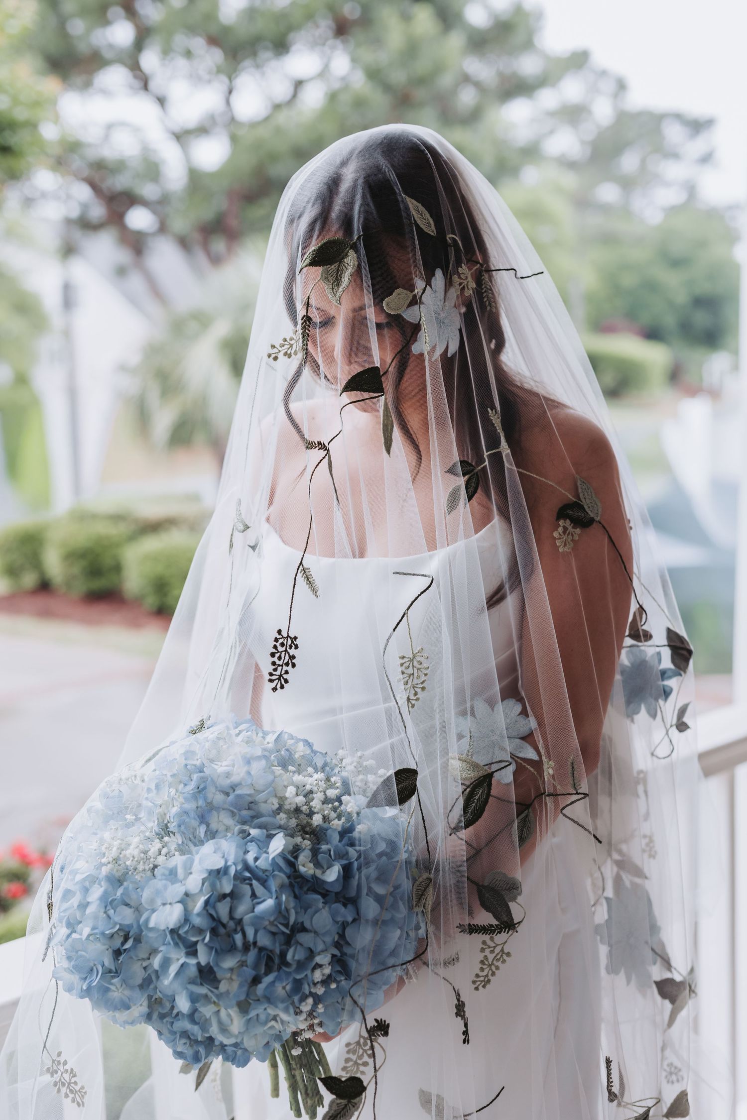 Bride in strapless gown holds blue hydrangea bouquet under floral embroidered veil outdoors.