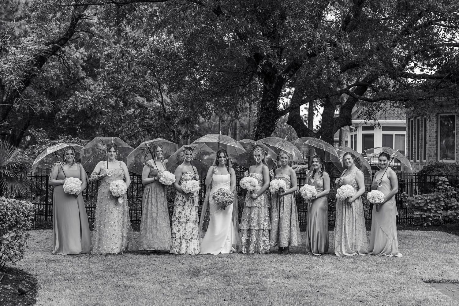 Bride and bridesmaids in mismatched gowns pose with clear umbrellas in black and white wedding photo.