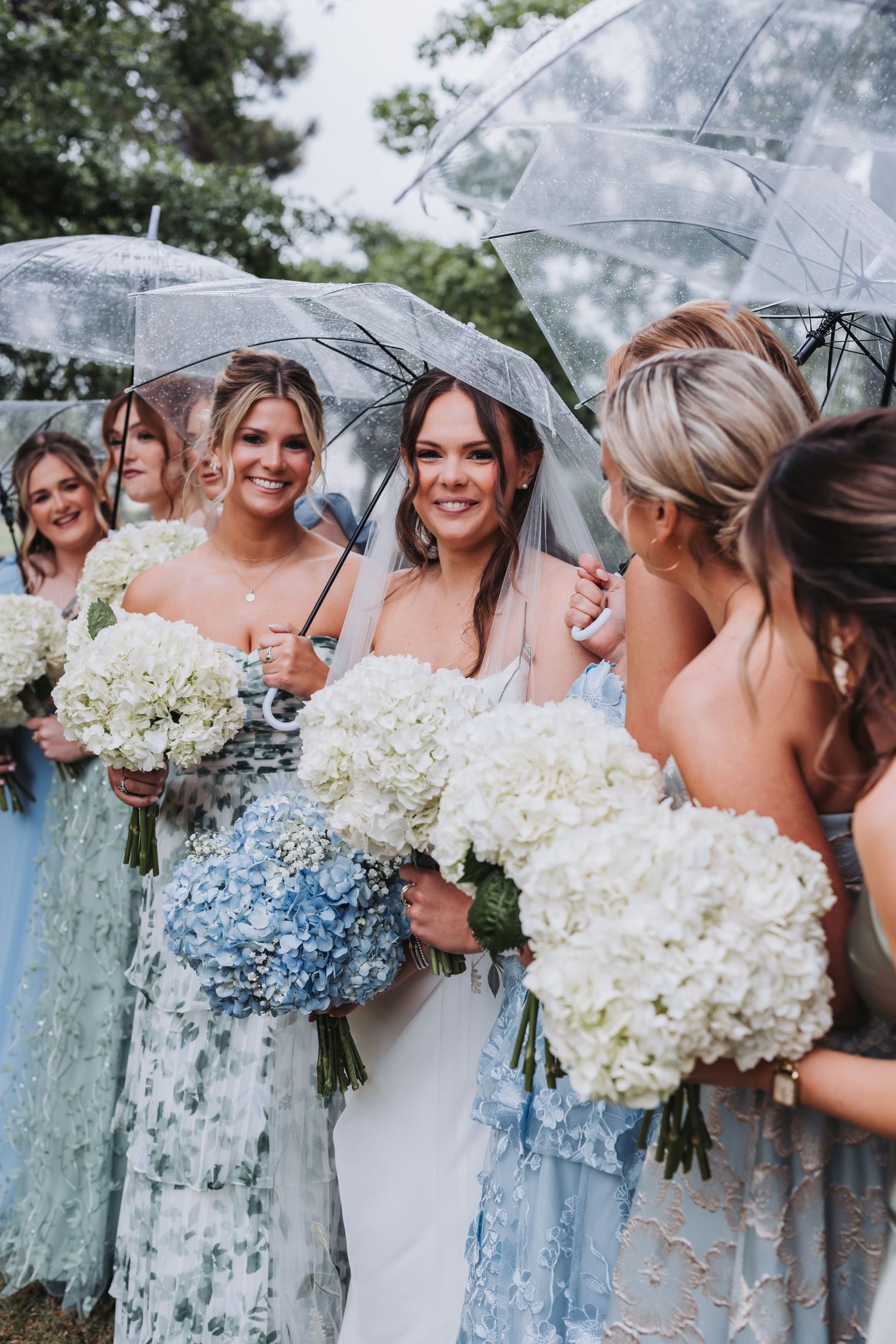 Smiling bride and bridesmaids in blue dresses holding white and blue hydrangea bouquets under clear umbrellas outdoors.