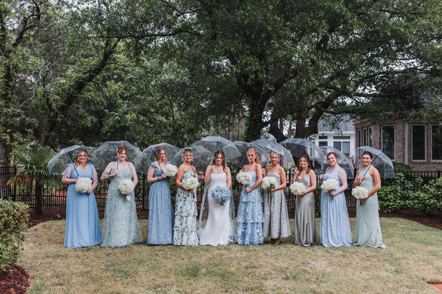 Bride and large bridal party in mismatched blue gowns holding bouquets under clear umbrellas on a rainy day.
