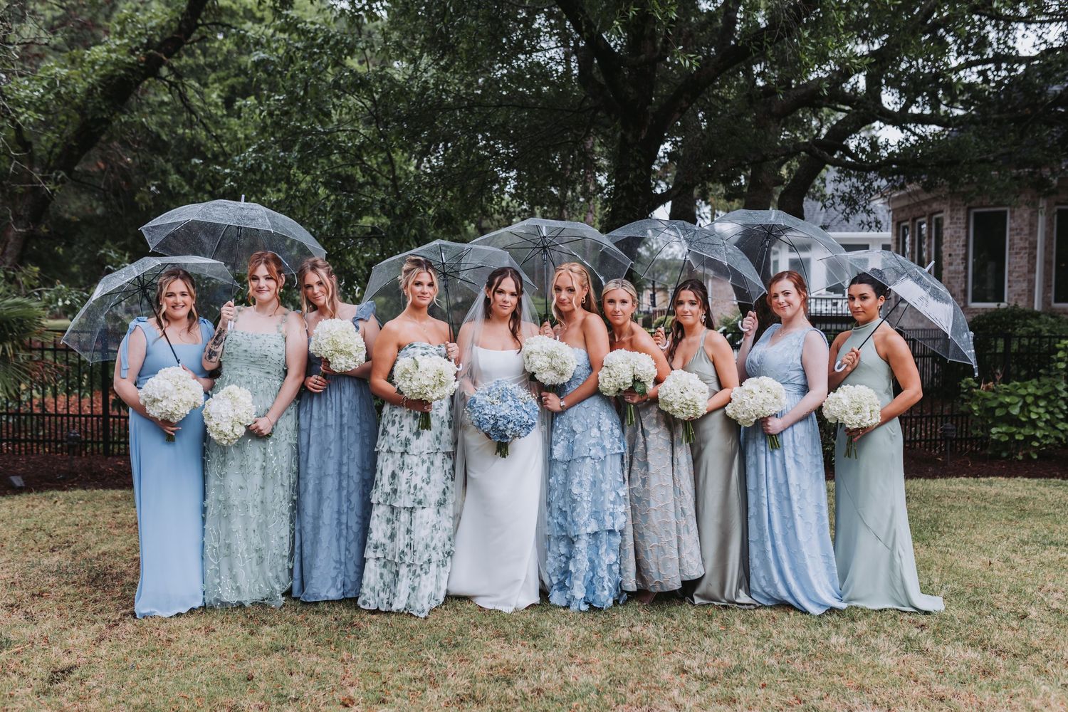 Bridal party in mismatched blue and sage gowns holding white bouquets under clear umbrellas on rainy wedding day.