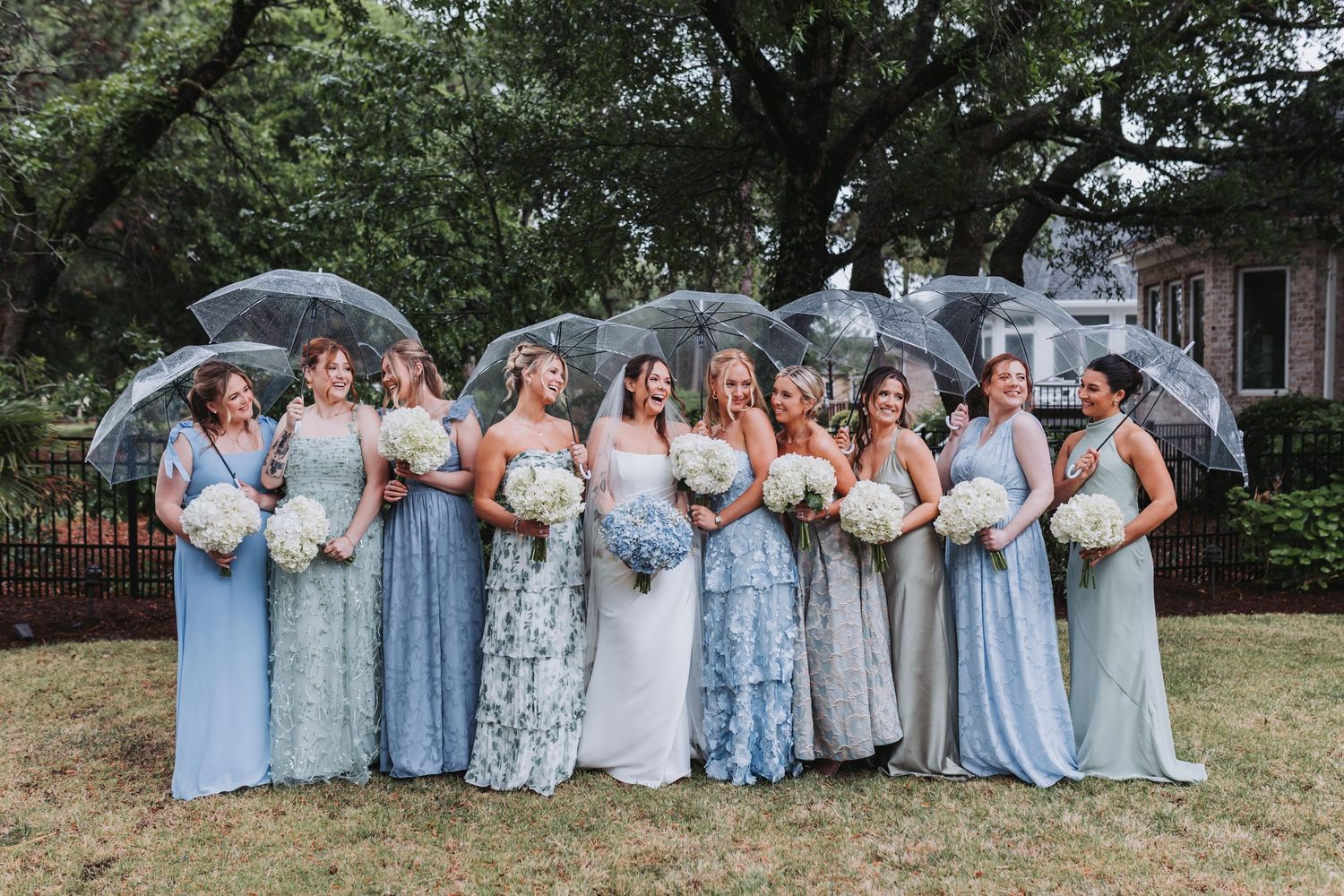 Bride surrounded by bridesmaids in various blue and sage gowns holding bouquets under clear umbrellas outdoors.