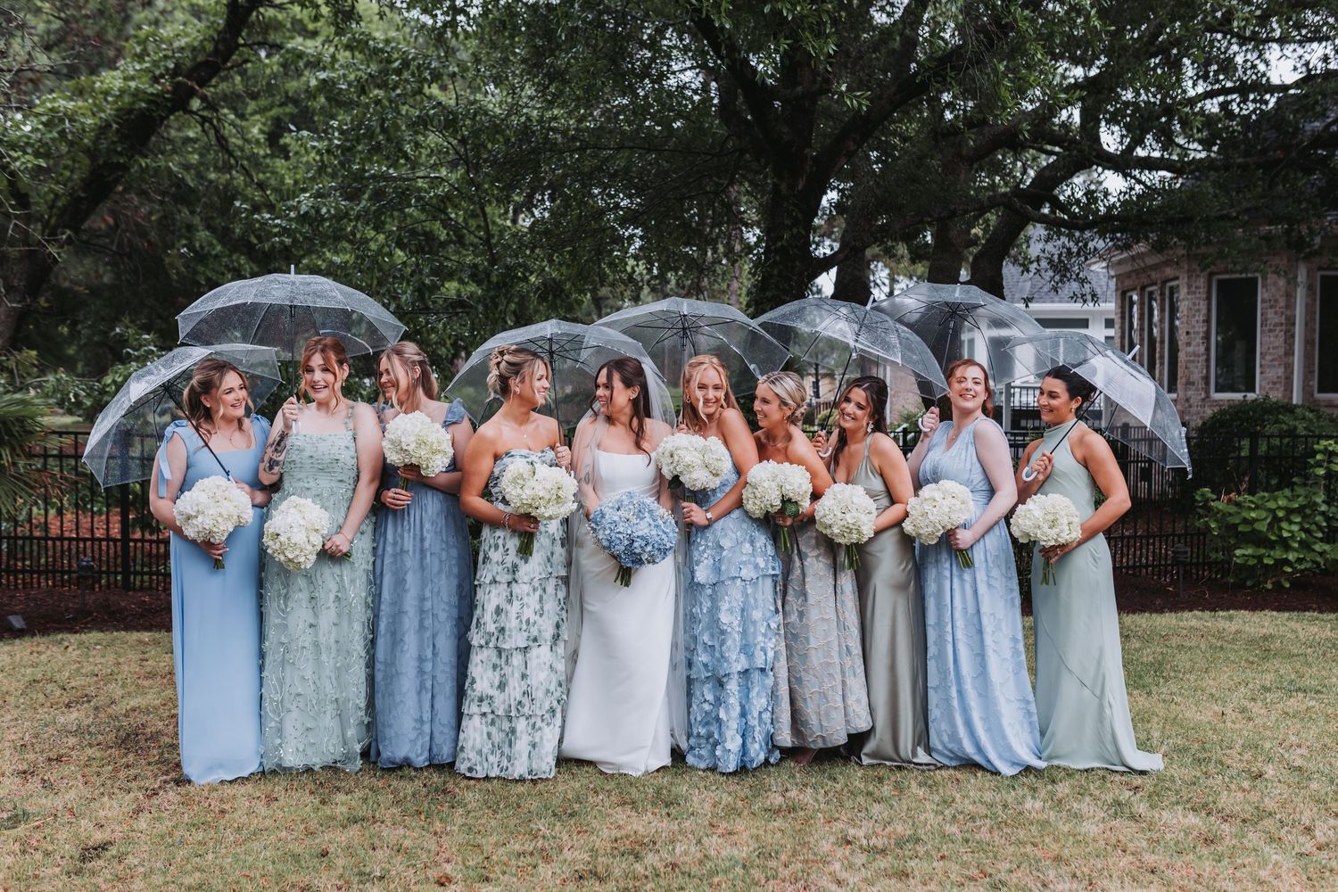 Bridal party in blue dresses holding clear umbrellas outdoors on rainy wedding day.