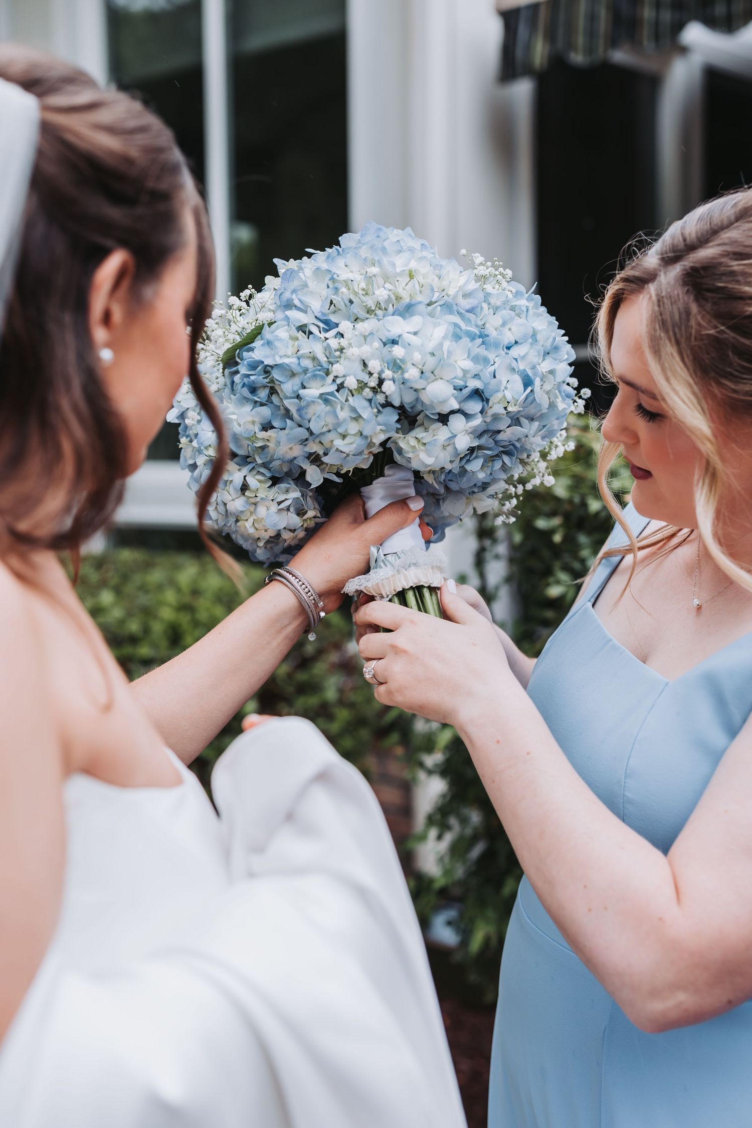 Bridesmaid in light blue dress adjusting ribbon on bride's blue hydrangea and baby's breath wedding bouquet outdoors.