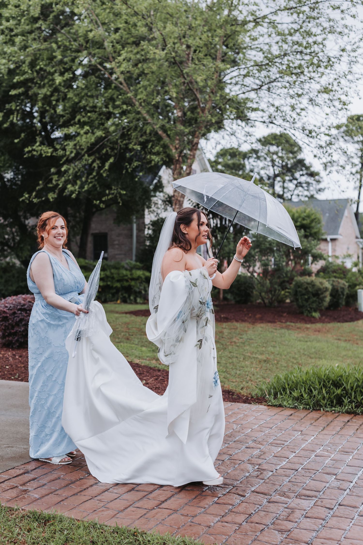 Bride holding clear umbrella in floral veil while bridesmaid carries her white gown train on a rainy wedding day.