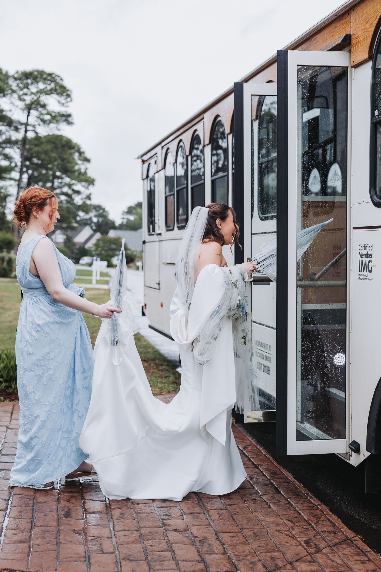 Bridesmaid in light blue gown helps bride in white dress and veil board a white trolley on wedding day.