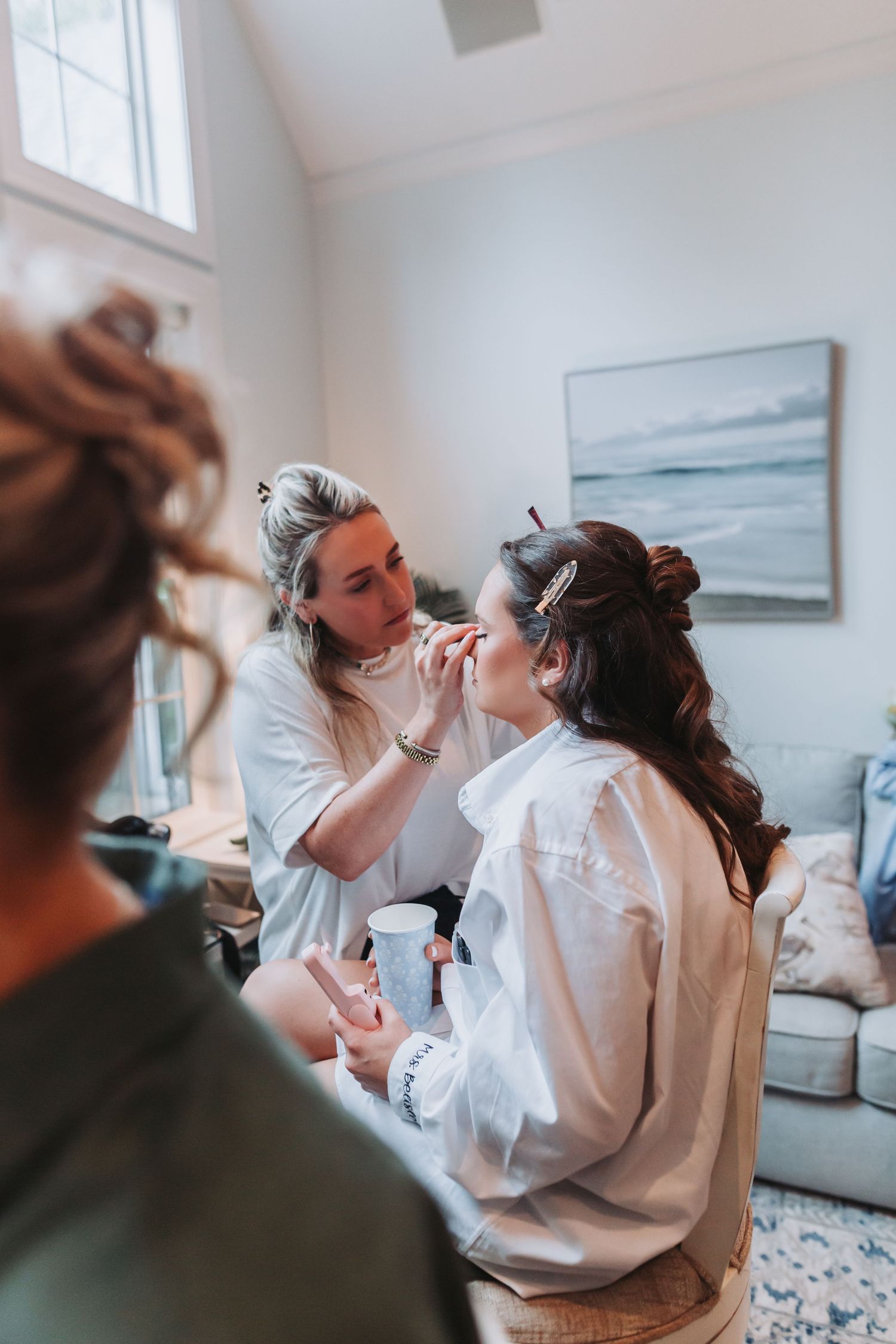 Makeup artist applying cosmetics to bride in white robe during wedding preparation in elegant room.