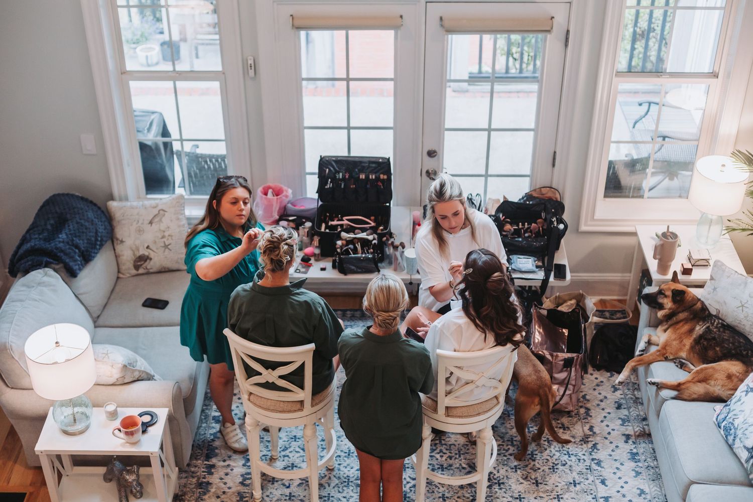 Bridal party getting hair and makeup done in a bright sunlit living room with a dog resting nearby.