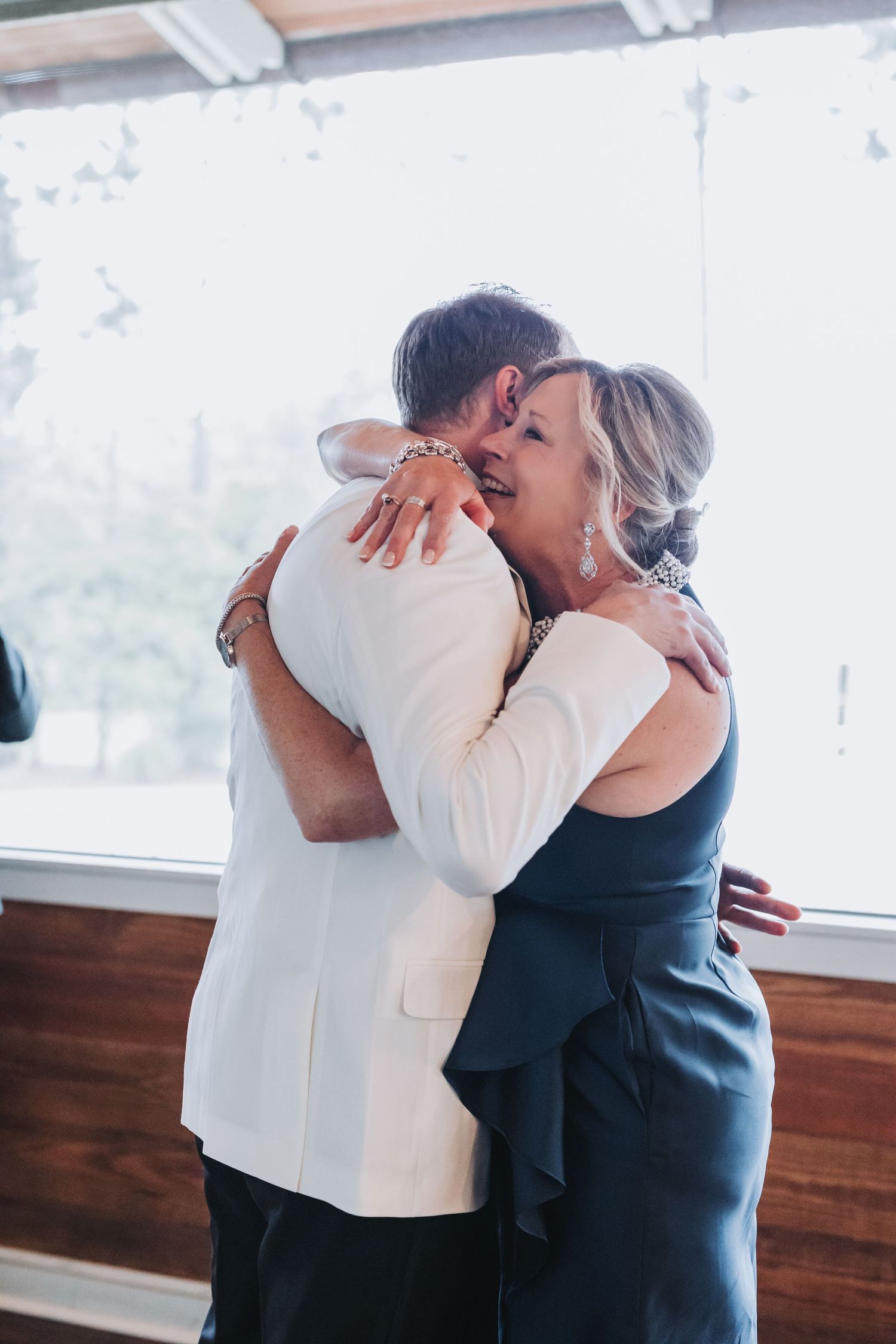 Groom in white tuxedo sharing an emotional hug with a woman in a blue dress.