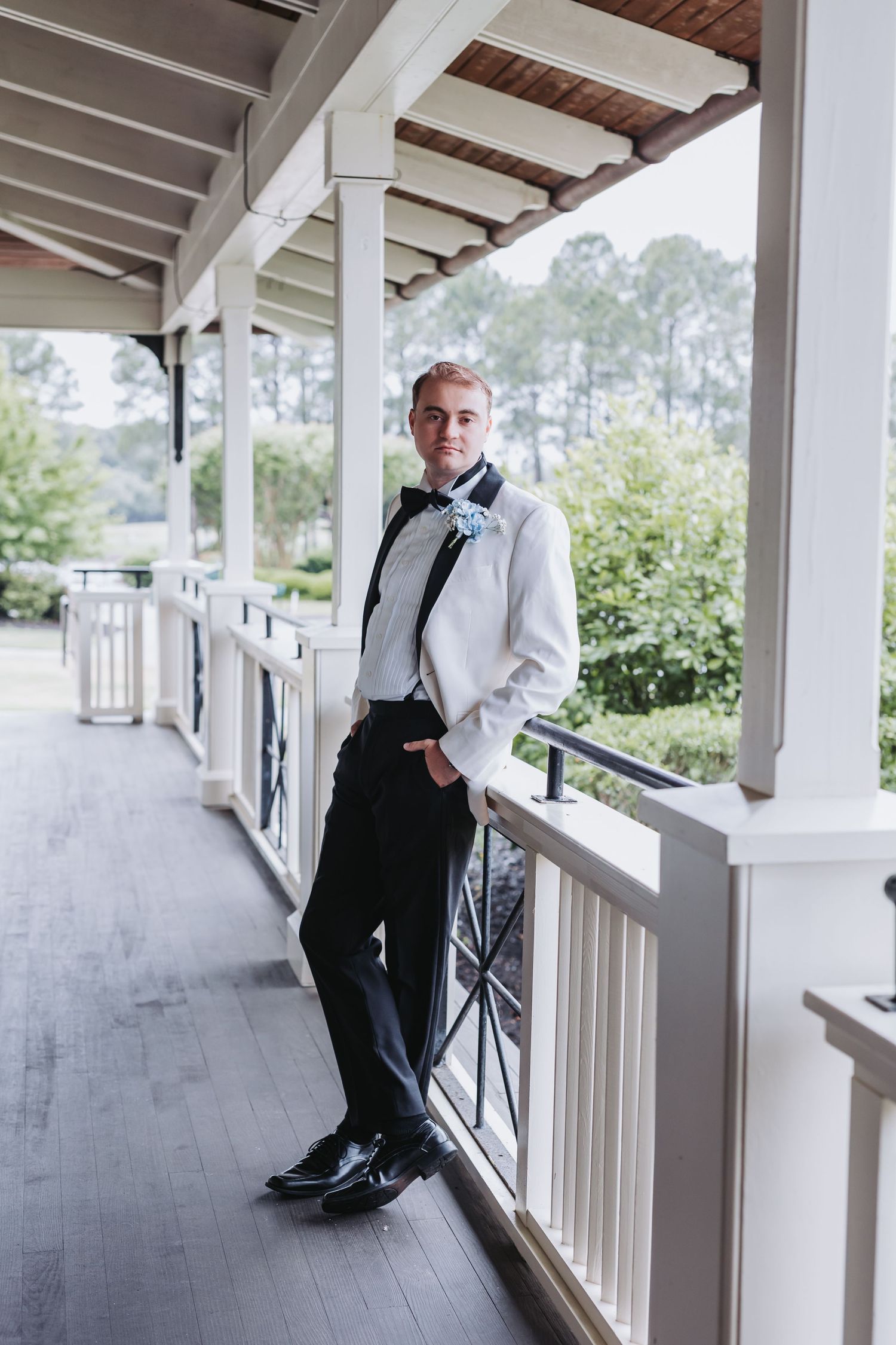 Dapper groom in white tuxedo jacket and black bow tie poses confidently on elegant covered porch.