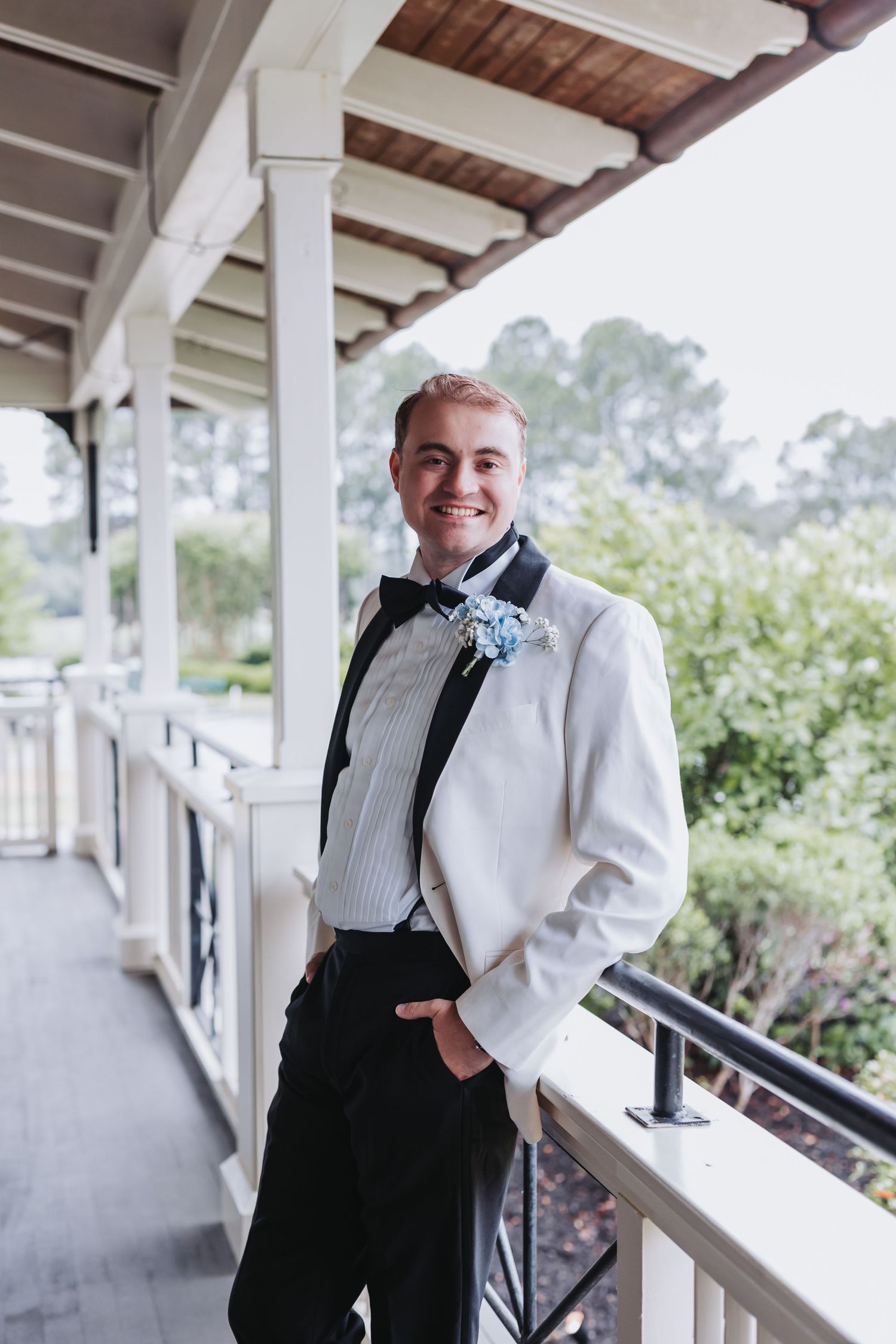 Smiling groom in white tuxedo jacket with blue boutonniere poses on elegant venue porch.
