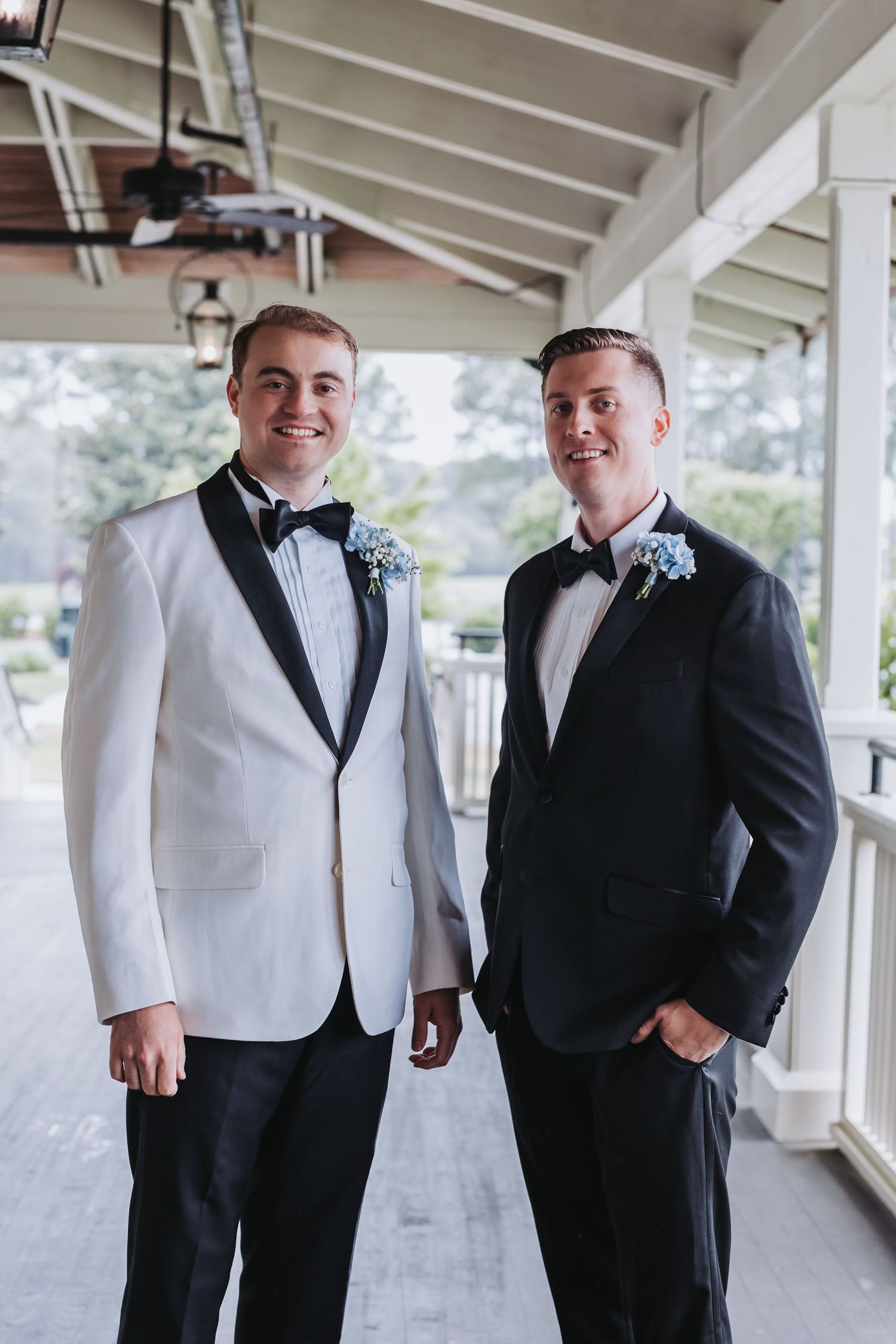 Two grooms in tuxedos with blue boutonnieres pose on a porch, one in white jacket and one in black suit.