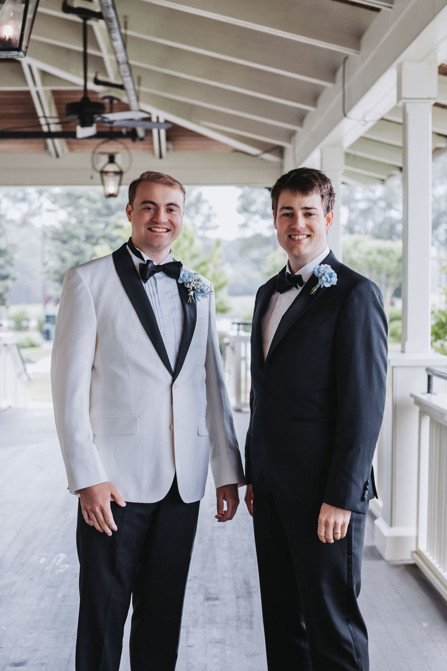 Two groomsmen in tuxedos with boutonnieres smiling on a covered porch at a wedding.