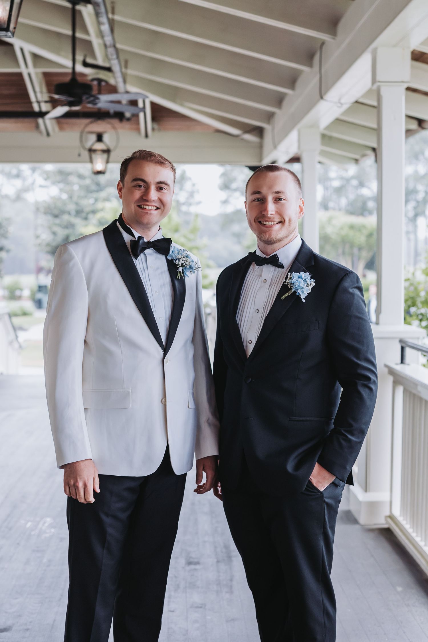 Two smiling groomsmen in black and white tuxedos with blue boutonnieres at outdoor wedding.