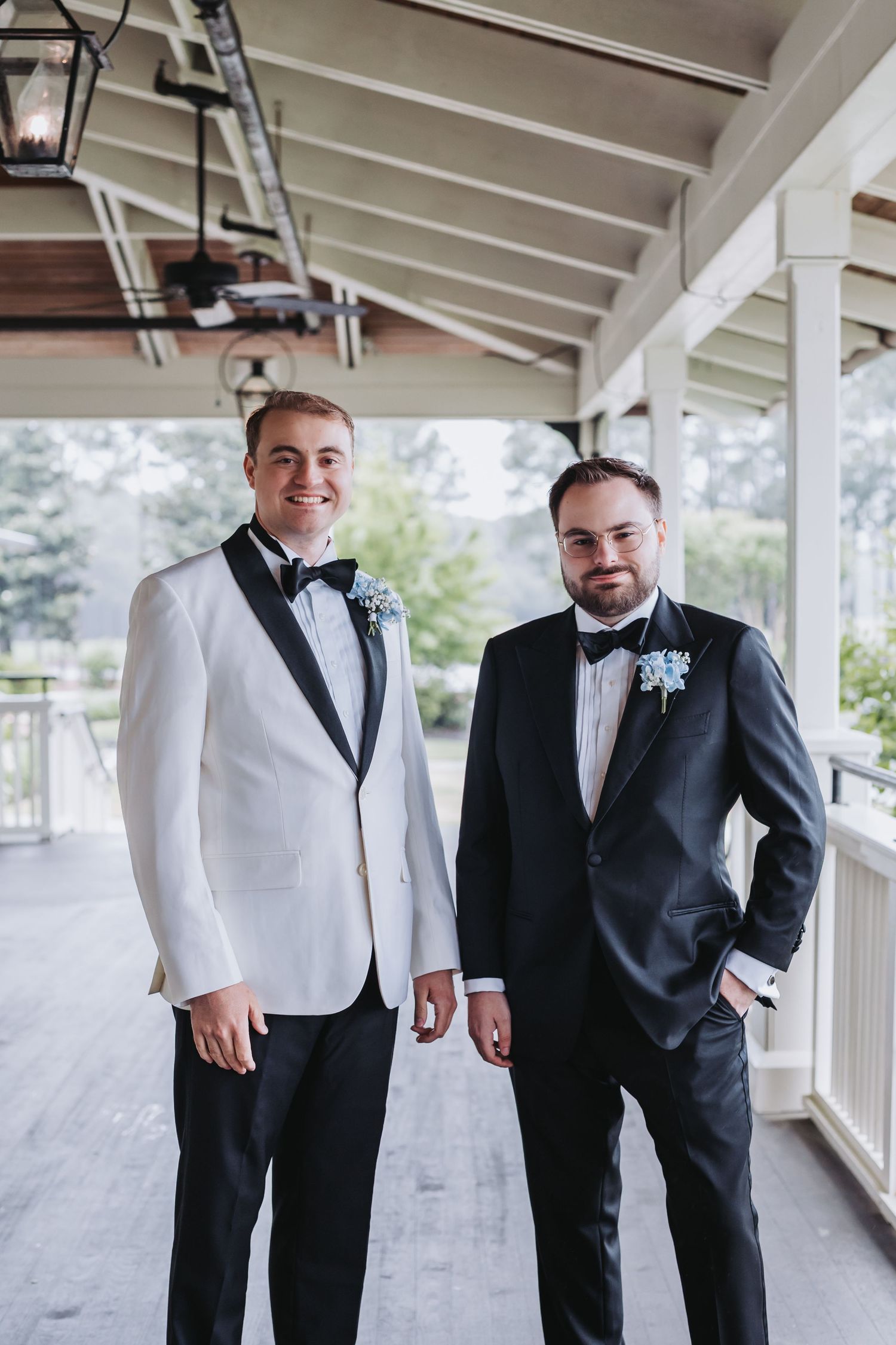 Groom in white tuxedo and groomsman in black suit pose together on wedding venue porch.