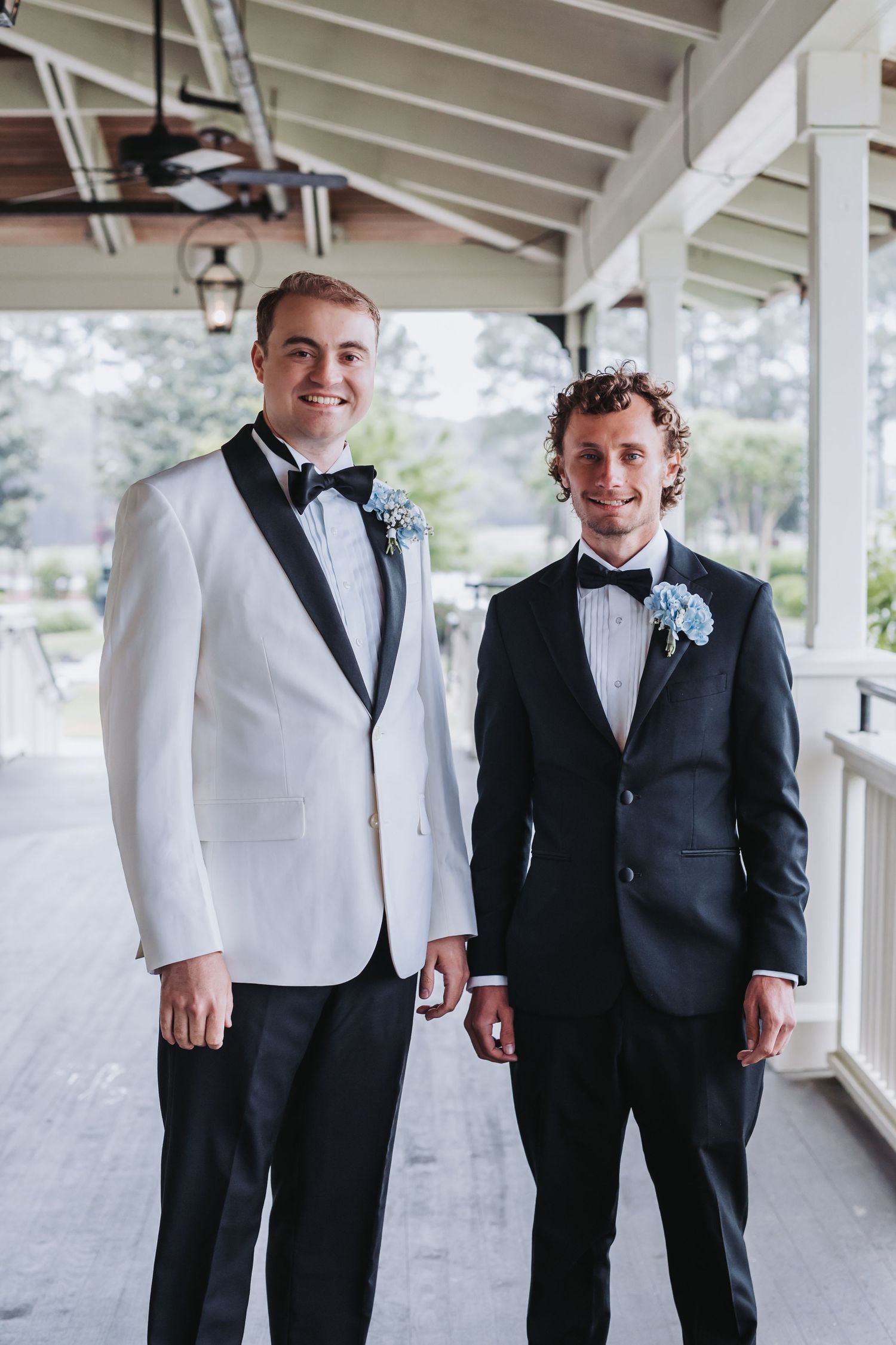Two grooms in tuxedos with boutonnieres smile on a covered porch at their wedding.
