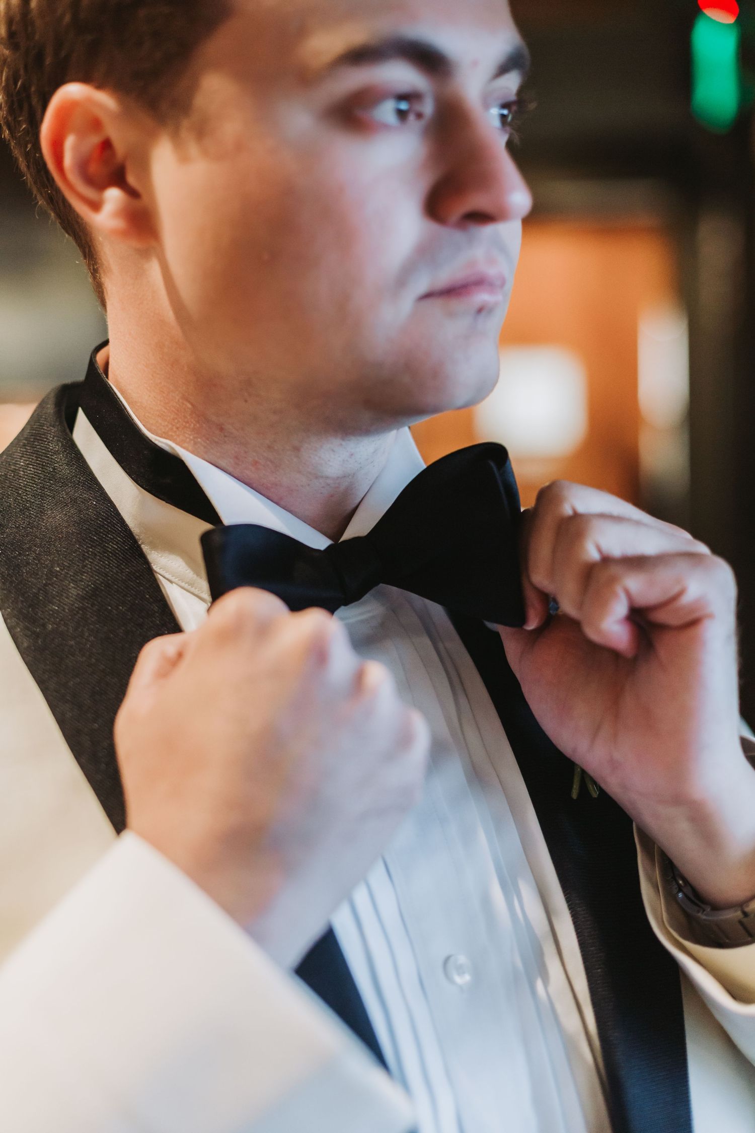 Groom in white tuxedo jacket adjusts black bow tie in close-up wedding portrait.