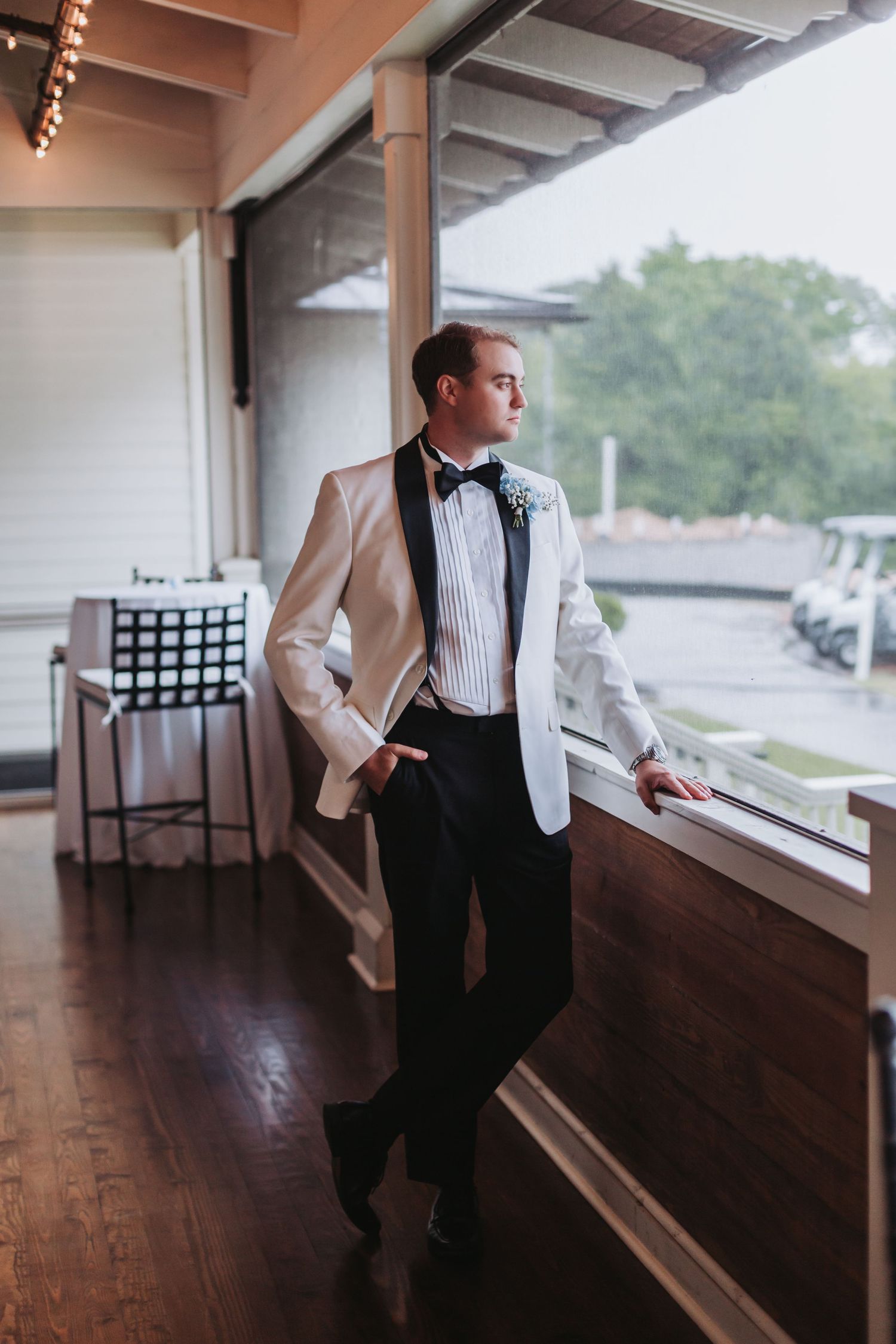 Stylish groom in white dinner jacket gazes thoughtfully out window at rainy wedding venue.