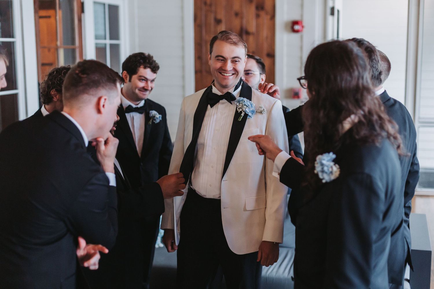 Groom in white tuxedo jacket laughing with groomsmen in black tuxedos before wedding ceremony.