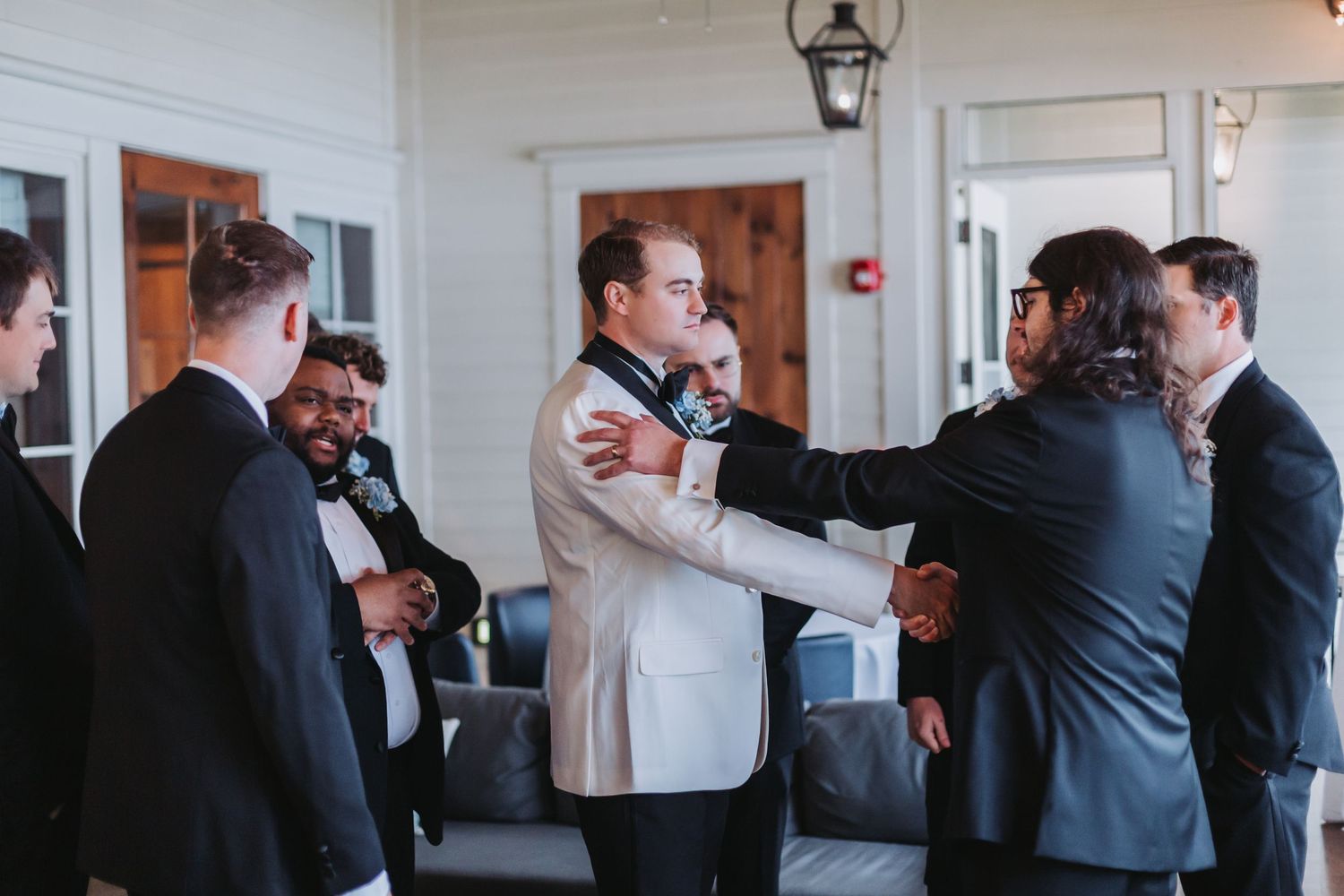 Groomsmen helping groom adjust his white tuxedo jacket before wedding ceremony indoors.