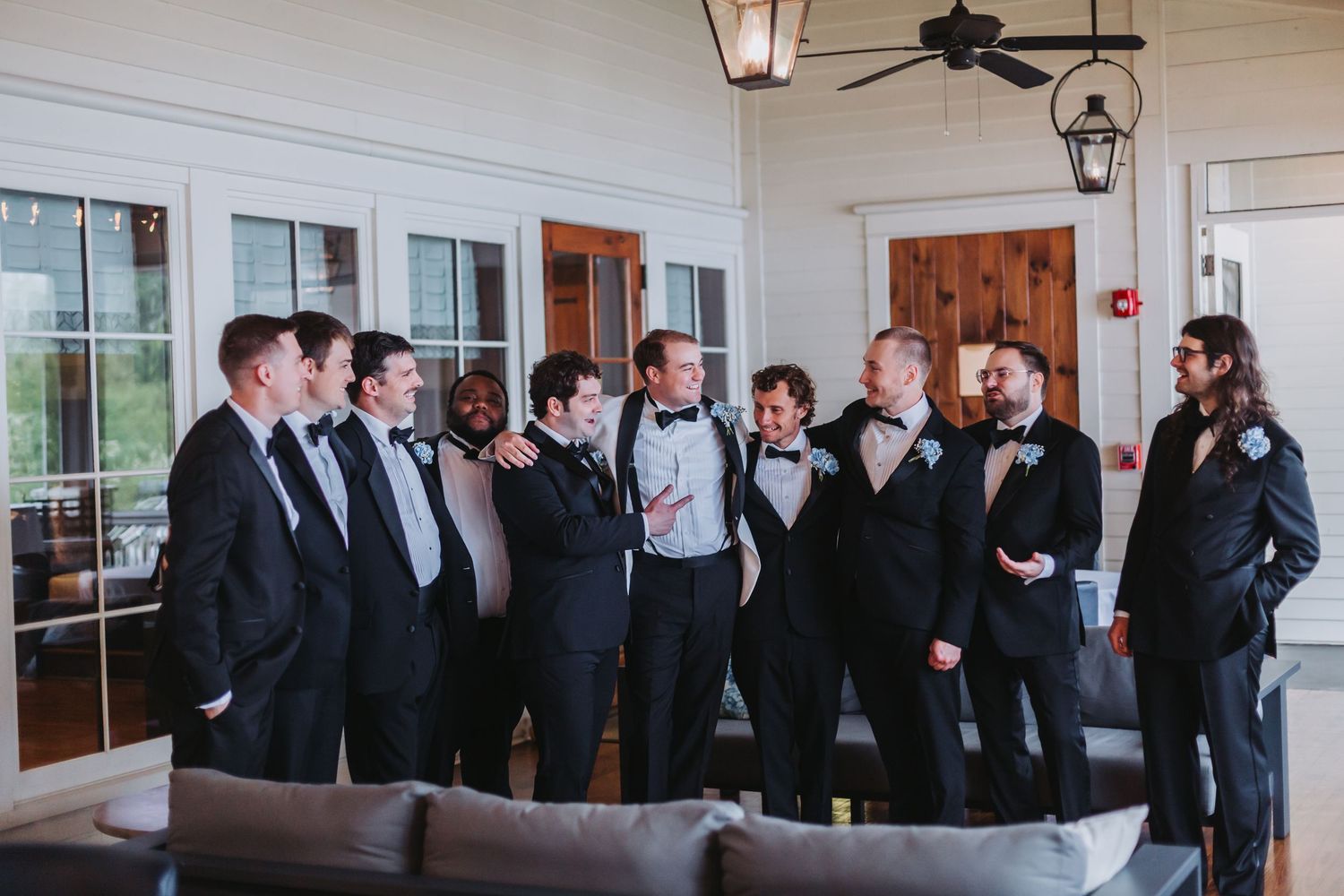 Groom and groomsmen in black tuxedos gathered together laughing in elegant venue interior.
