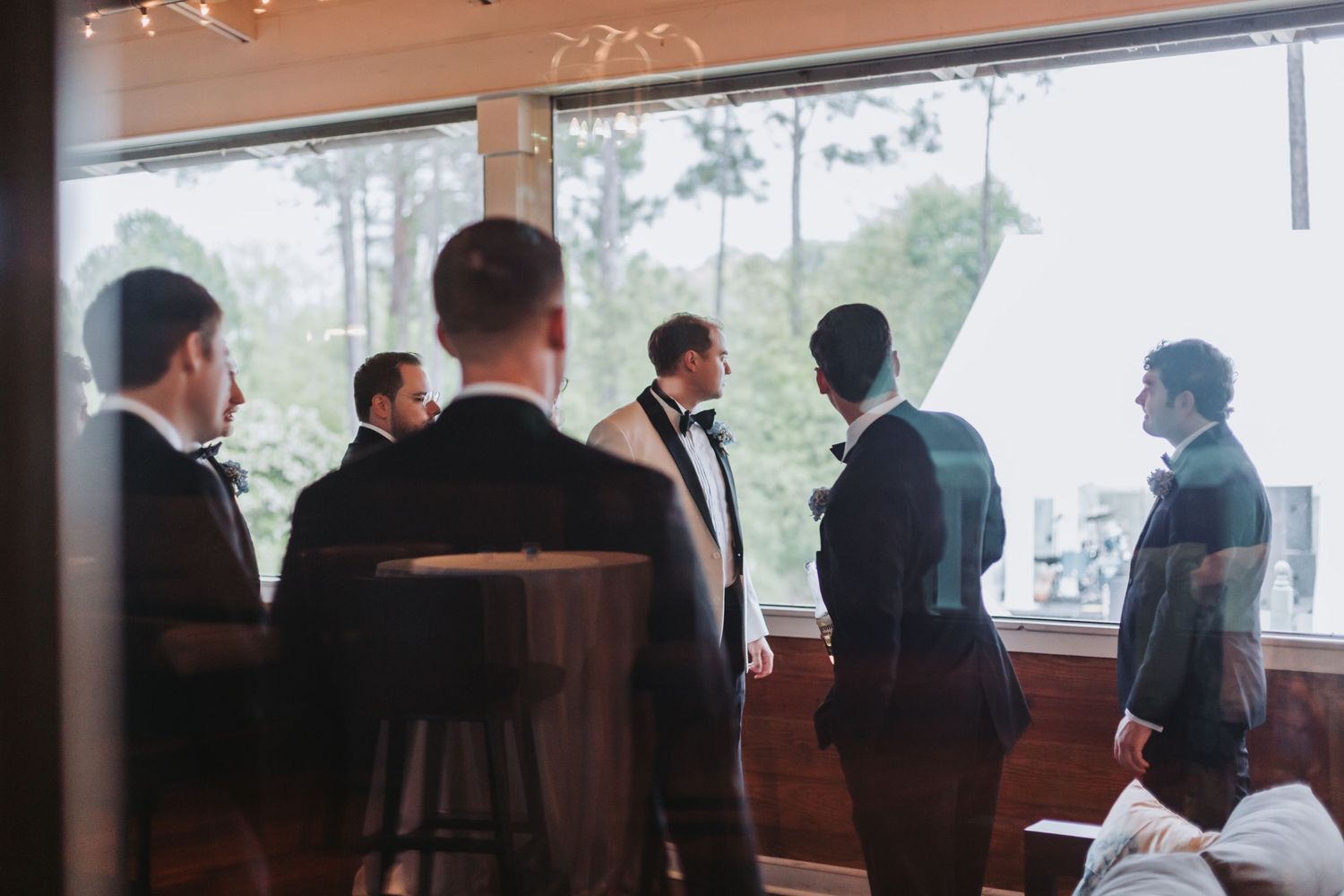 Groomsmen in black tuxedos gathered near large windows in elegant wedding venue.
