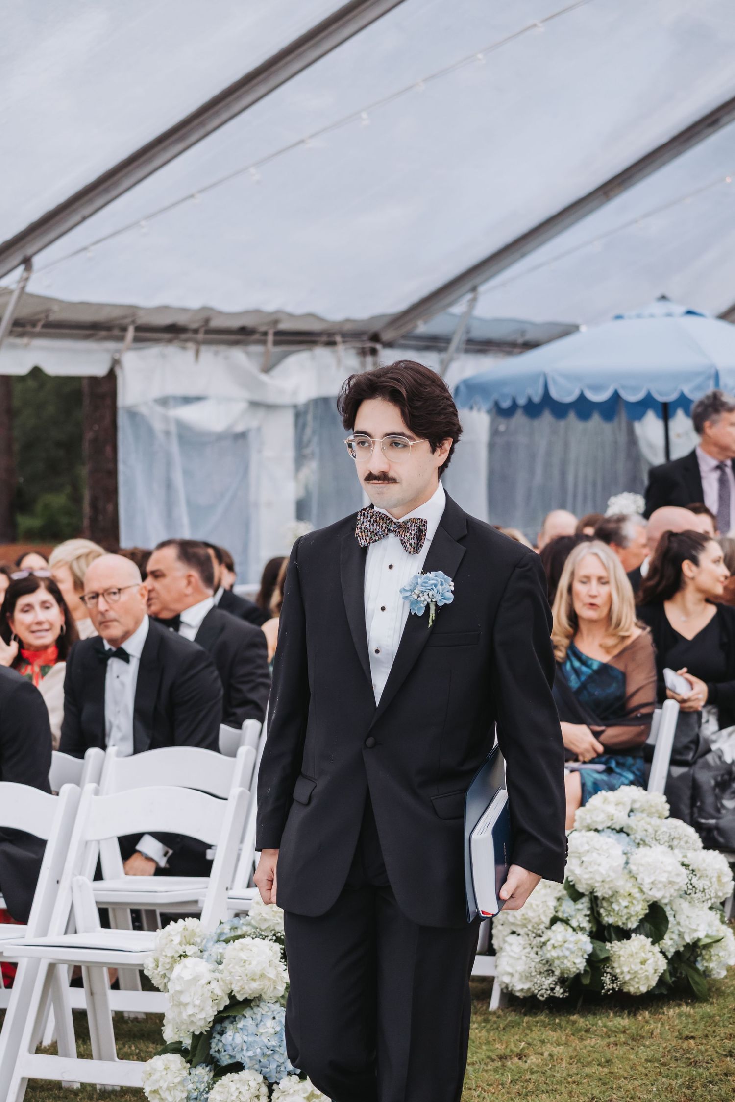 Groomsman in black tuxedo with mustache boutonniere walking down wedding aisle.