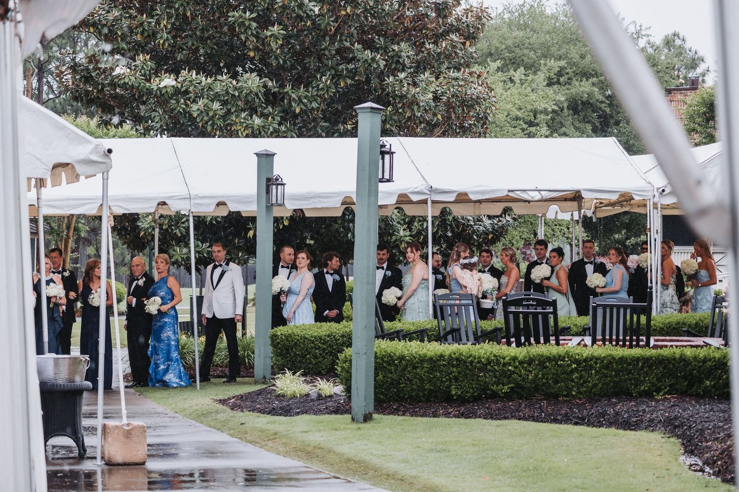 Wedding guests gather under white tent canopy during outdoor rainy day wedding ceremony on lush grounds.