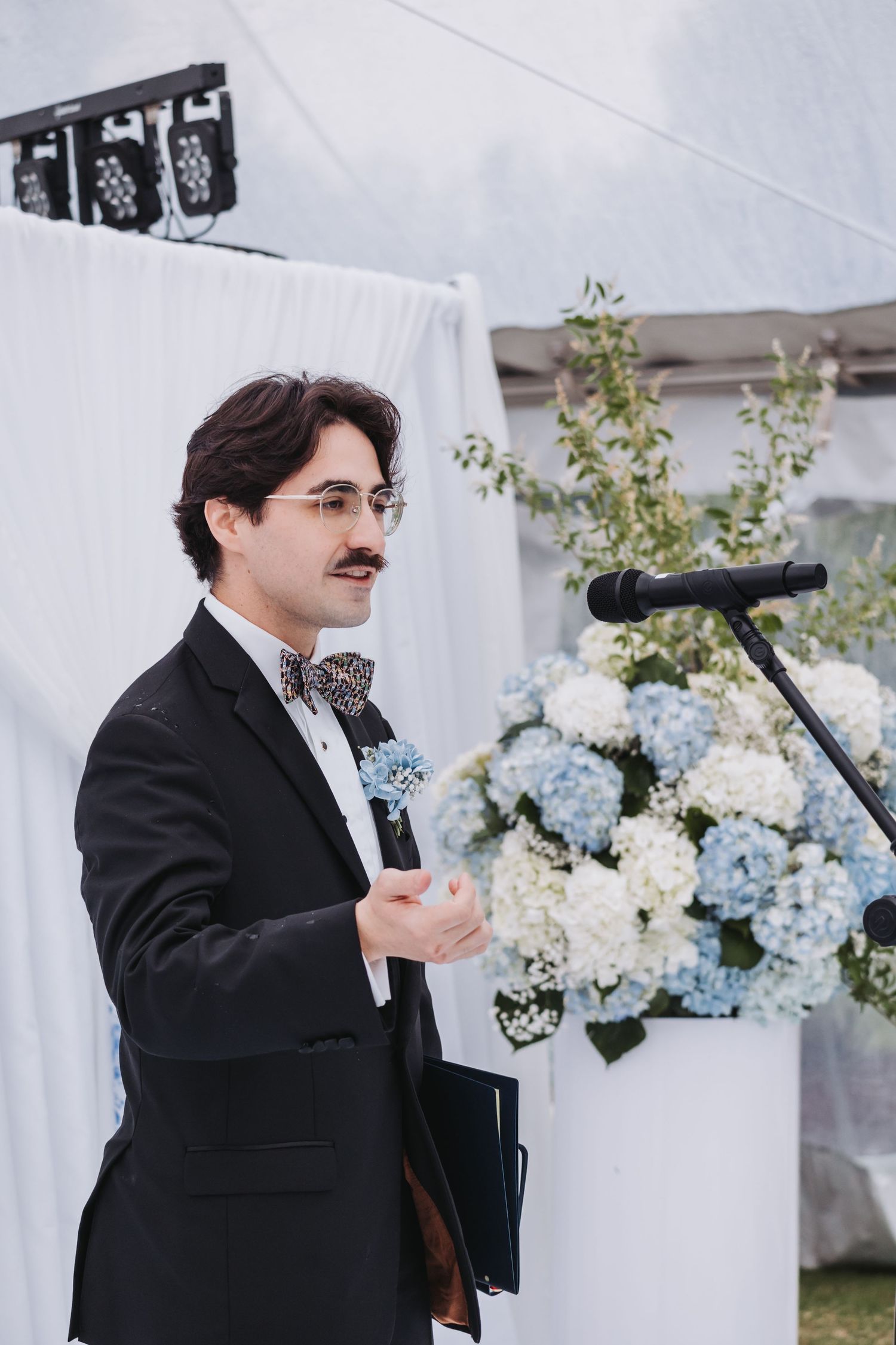 A dapper groom with a mustache speaks at a floral podium adorned with blue and white hydrangeas at an outdoor wedding.