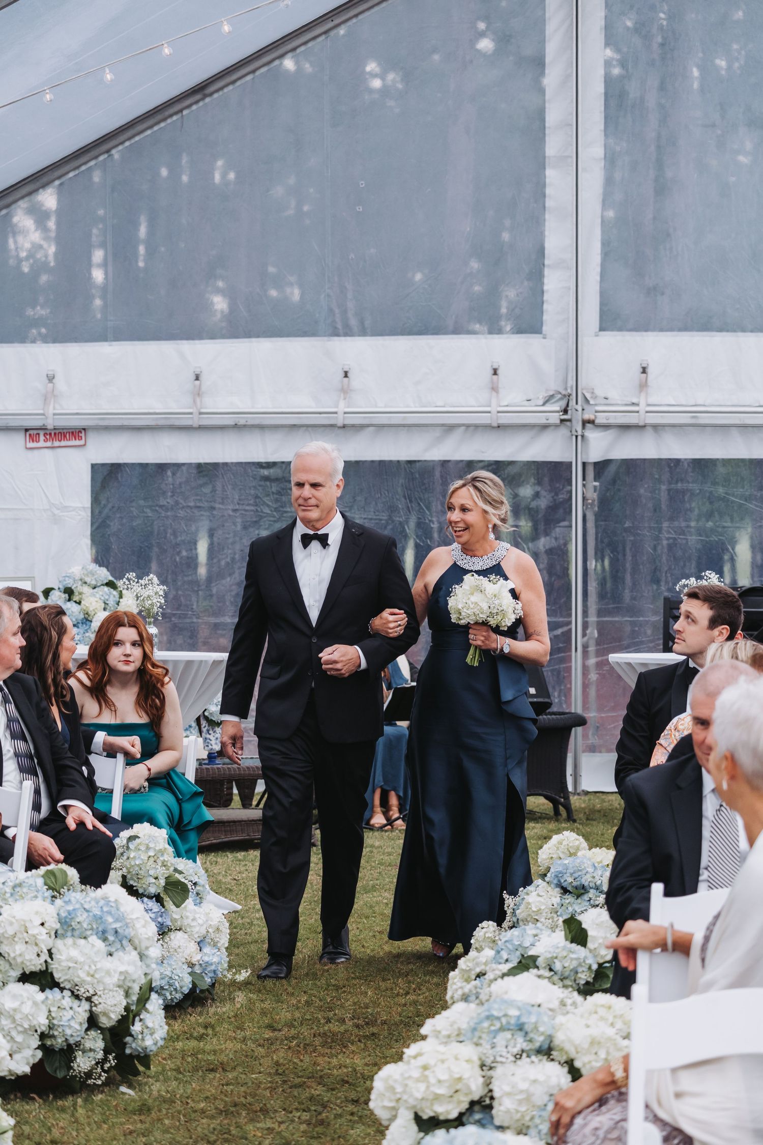 Elegantly dressed couple walking down wedding aisle lined with blue hydrangea floral arrangements.